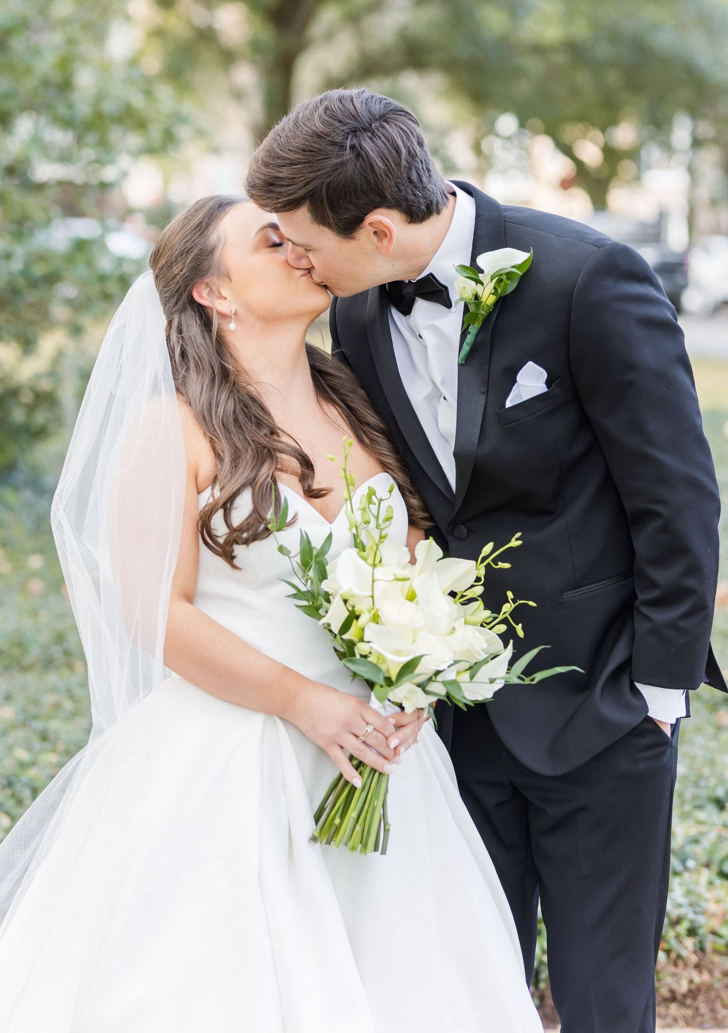 Bride and groom share a kiss outdoors, she holds white bouquet in wedding gown with veil, he wears black tuxedo.