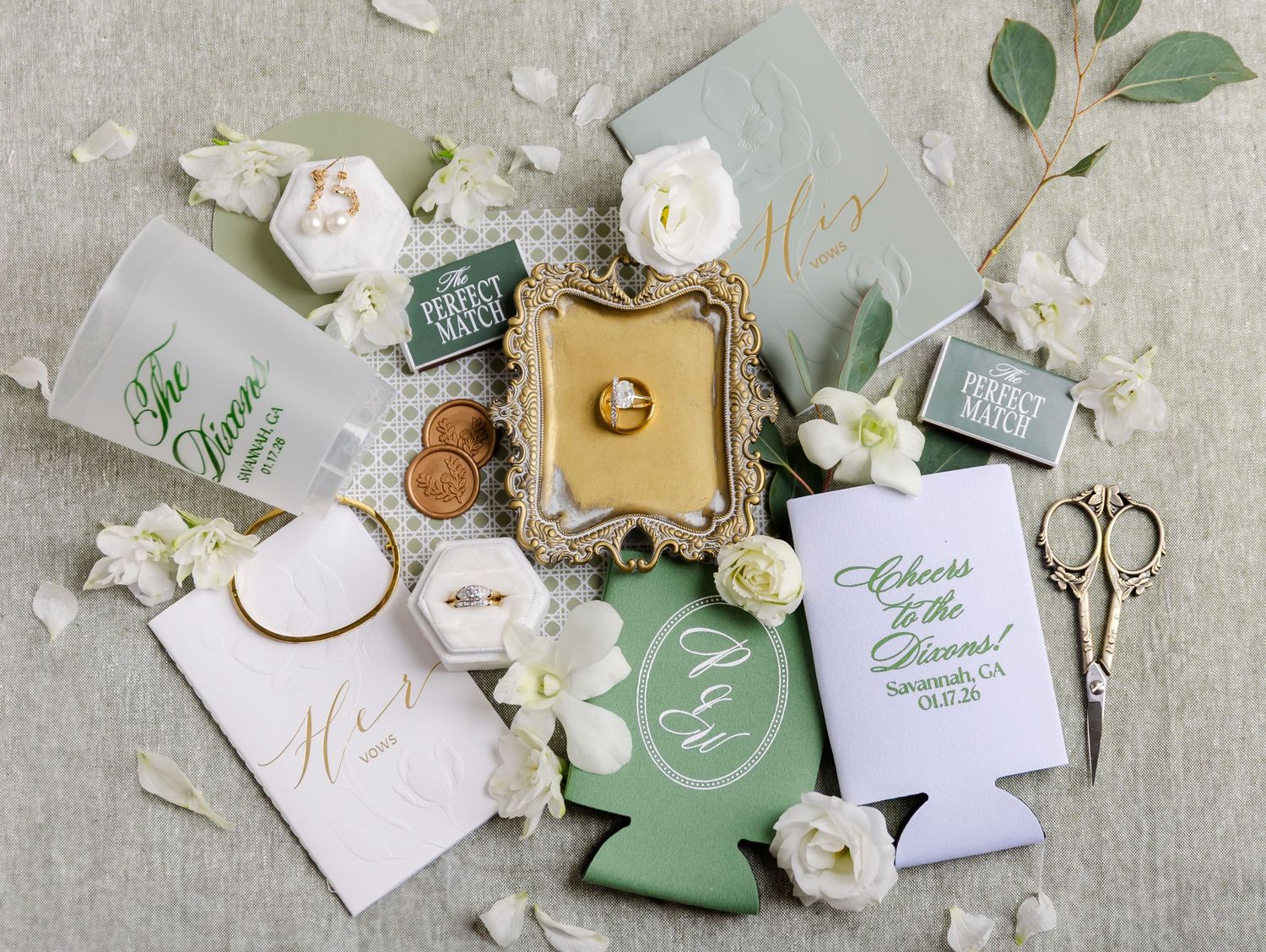 Wedding flat lay with sage green stationery, white floral details, gold ring dish, and personalized koozies.