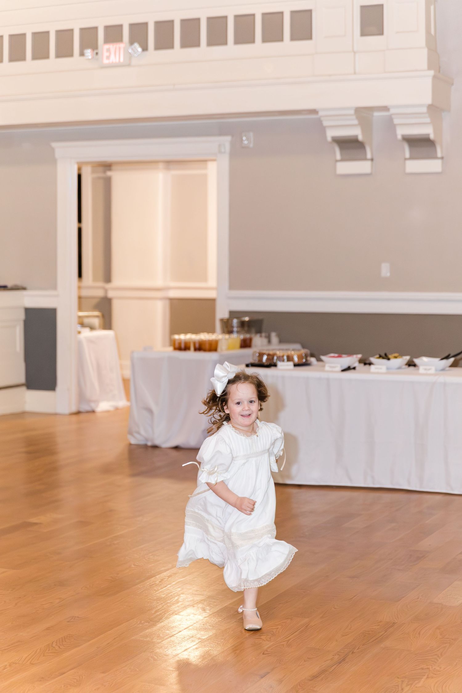 Joyful flower girl in white dress running across wooden dance floor at wedding reception.