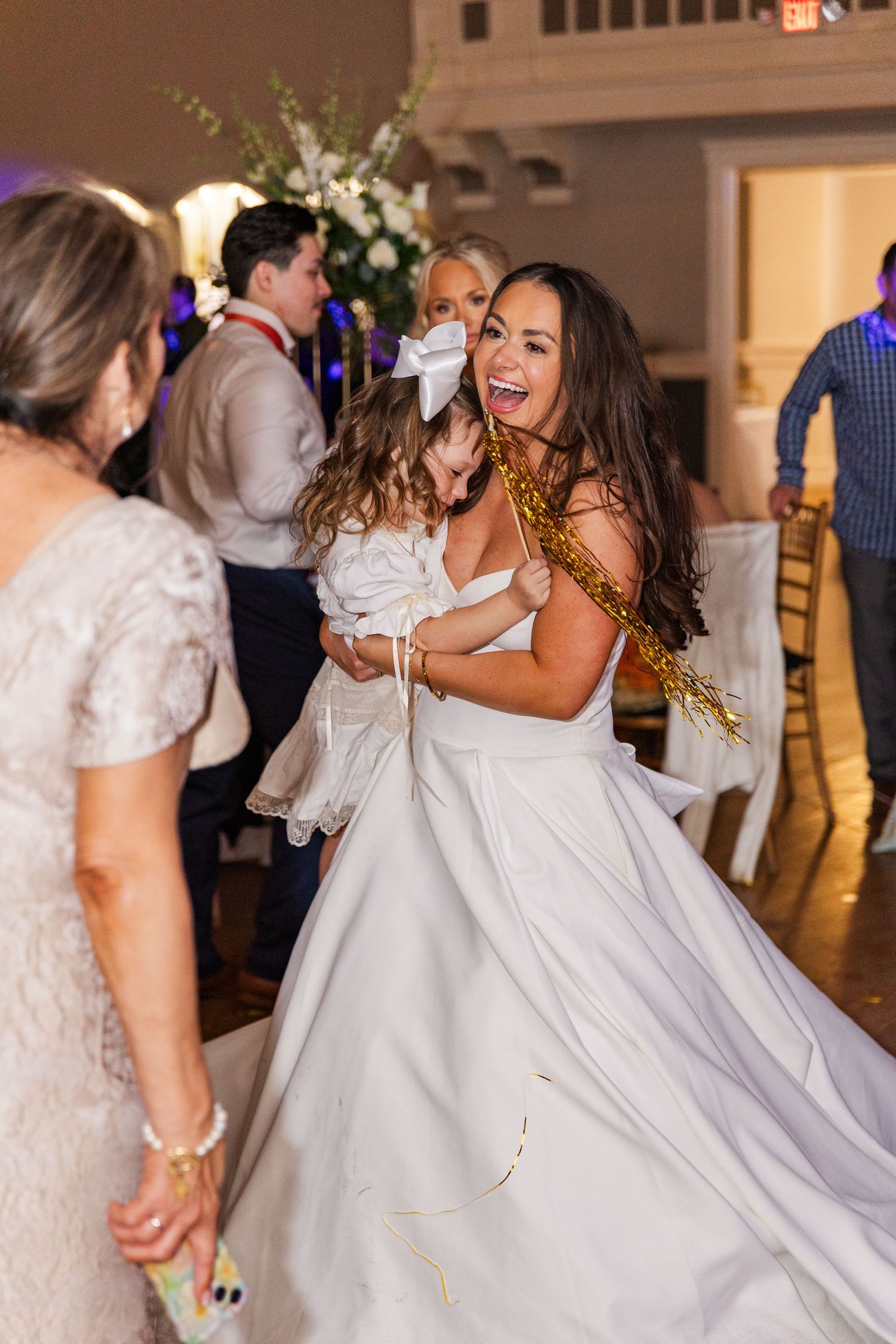 Joyful bride hugs flower girl while dancing at wedding reception holding gold tinsel streamers.