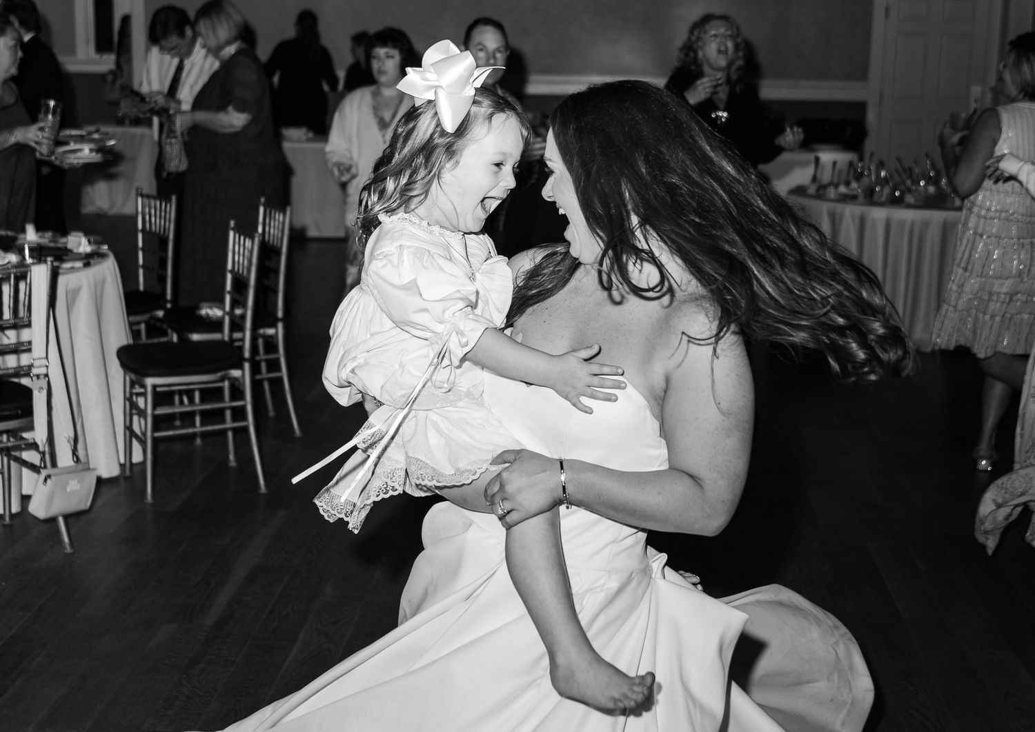 Black and white photo of a bride in a white gown holding a flower girl on a wedding reception dance floor.