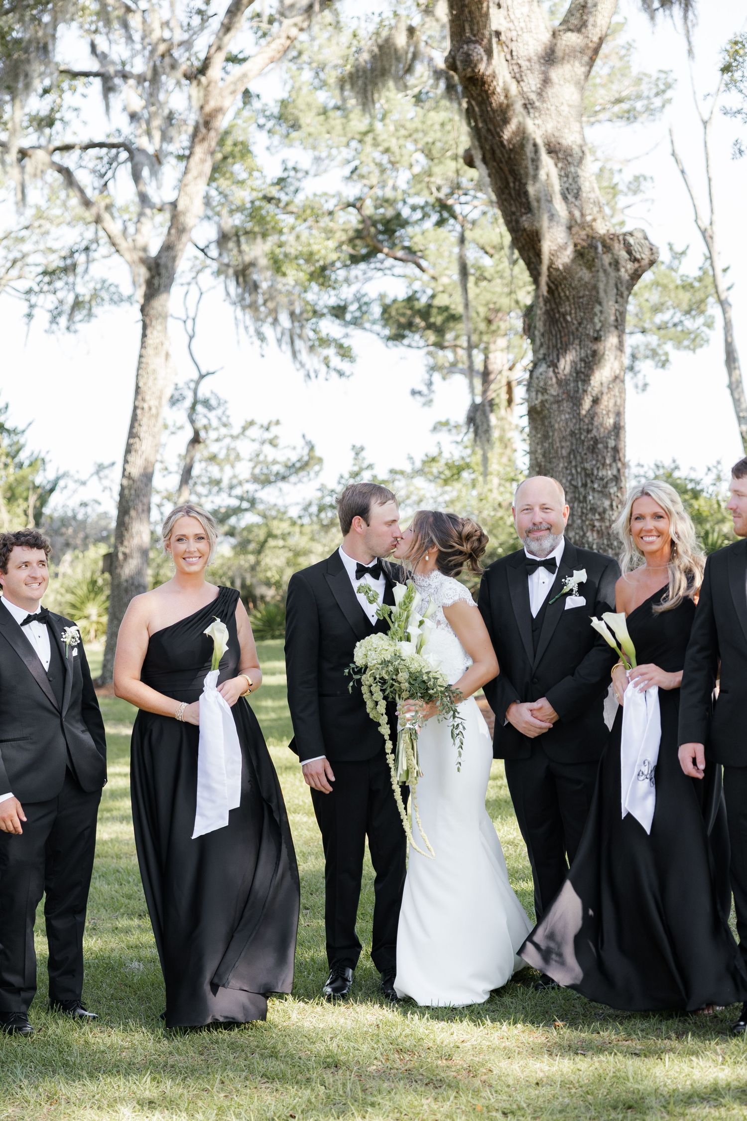 Wedding party in black attire poses outdoors as bride and groom share a kiss under moss-draped trees.