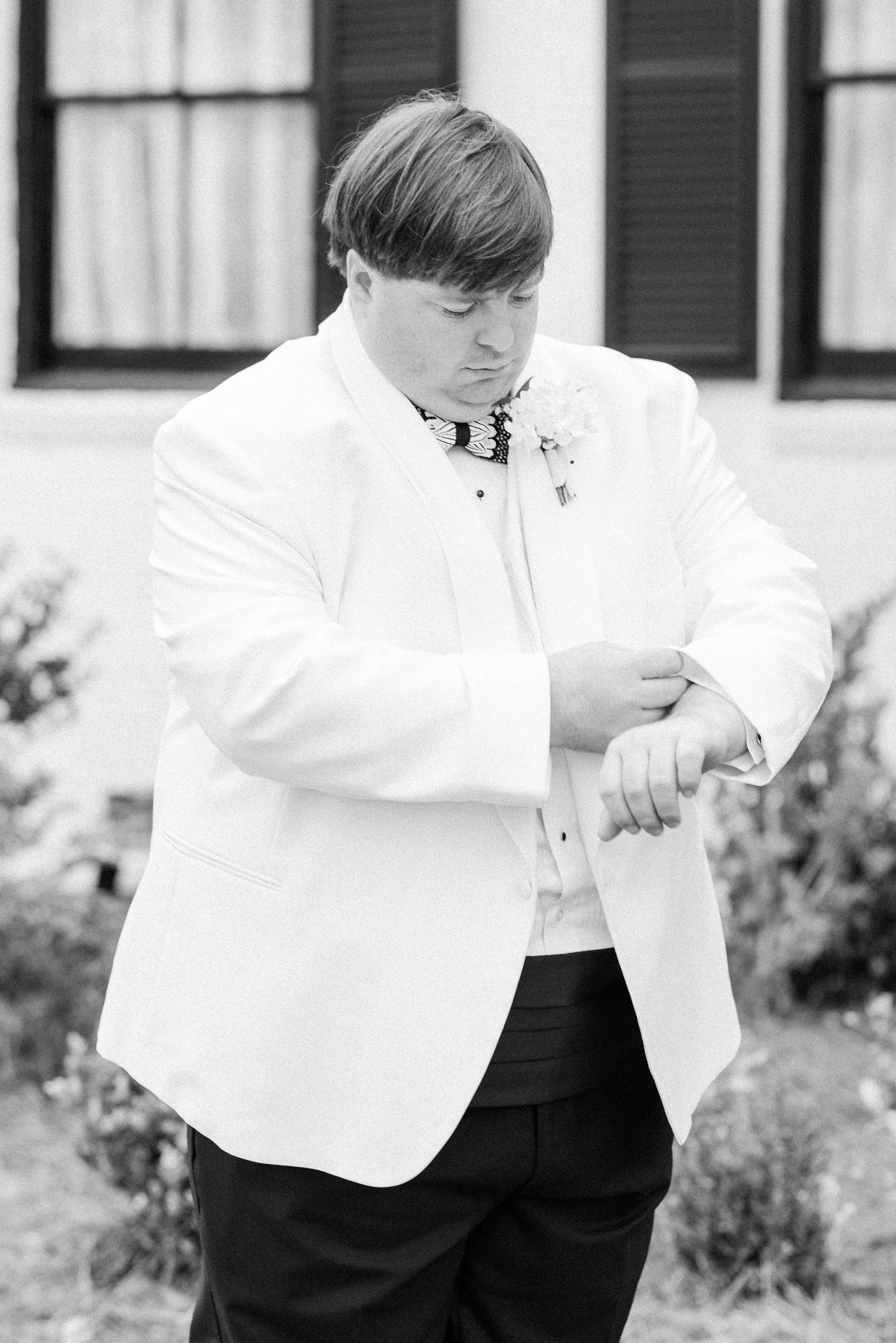 Groom in white tuxedo jacket checks his watch in black and white wedding portrait outside historic venue.