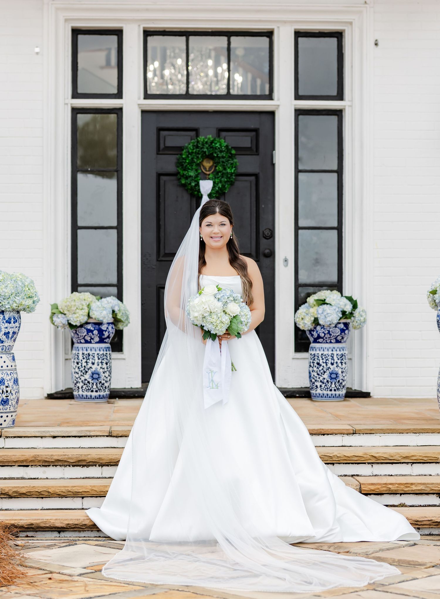 Bride in strapless white ball gown with cathedral veil poses before a white venue entrance with blue hydrangea urns.