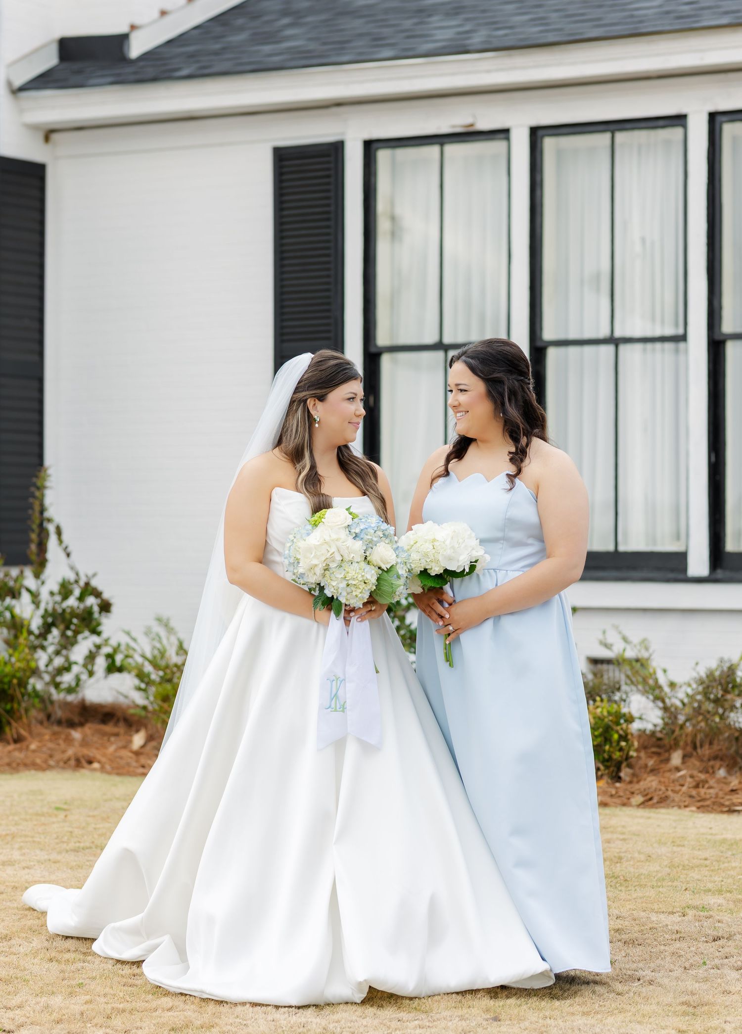 Bride in white strapless gown and bridesmaid in light blue dress smile at each other outside a white venue.