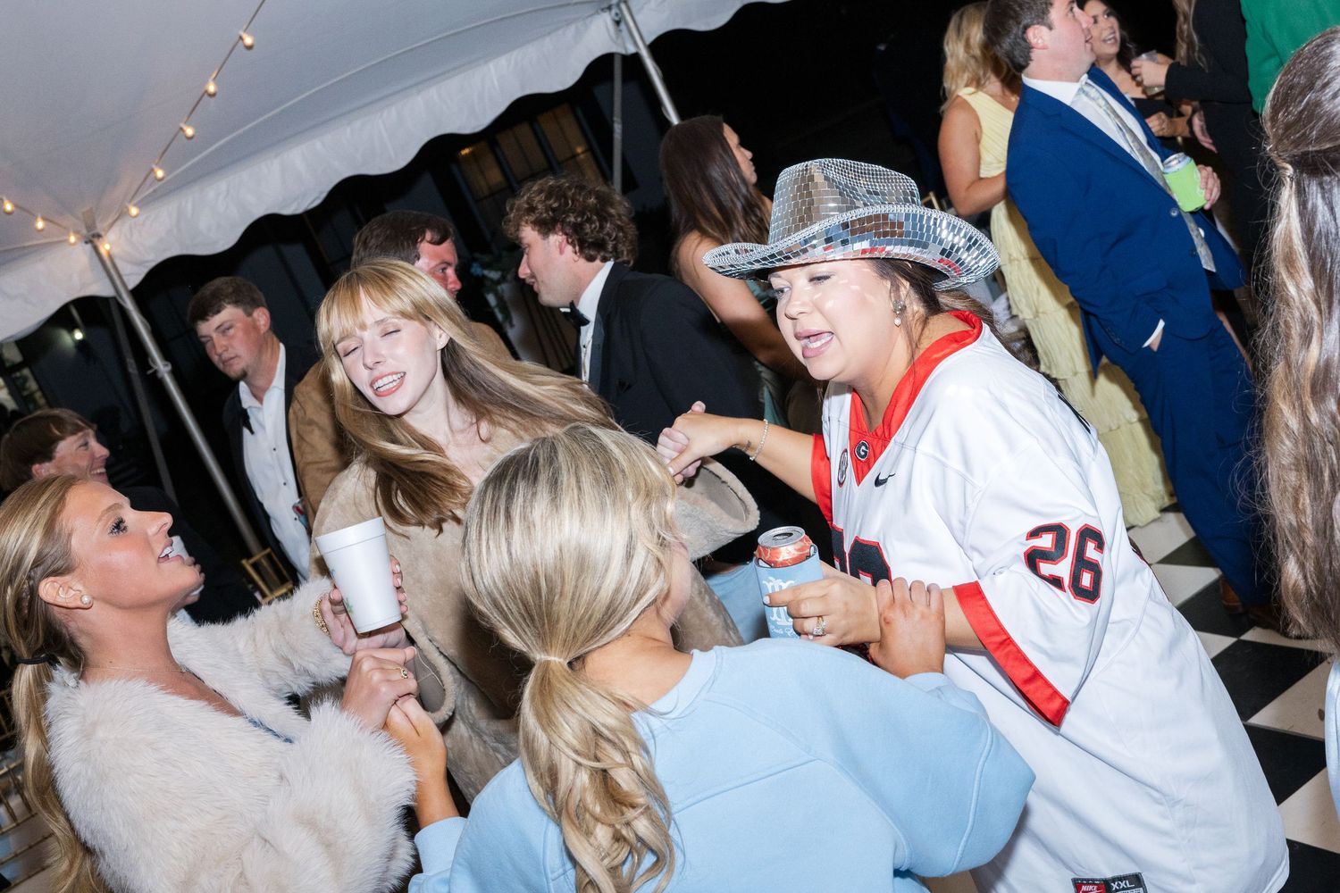 Wedding reception guests dancing under tent with woman in football jersey and silver cowboy hat at celebration.