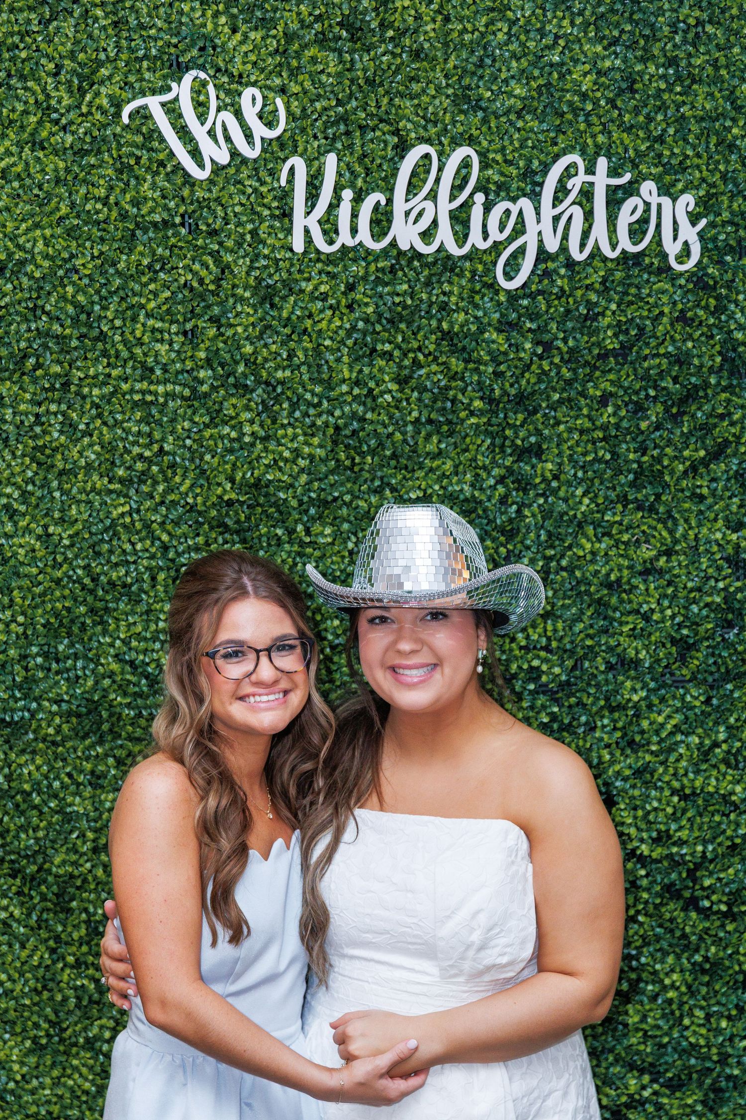 Two women pose in front of The Kicklighters green hedge sign wall at a wedding reception photo booth.