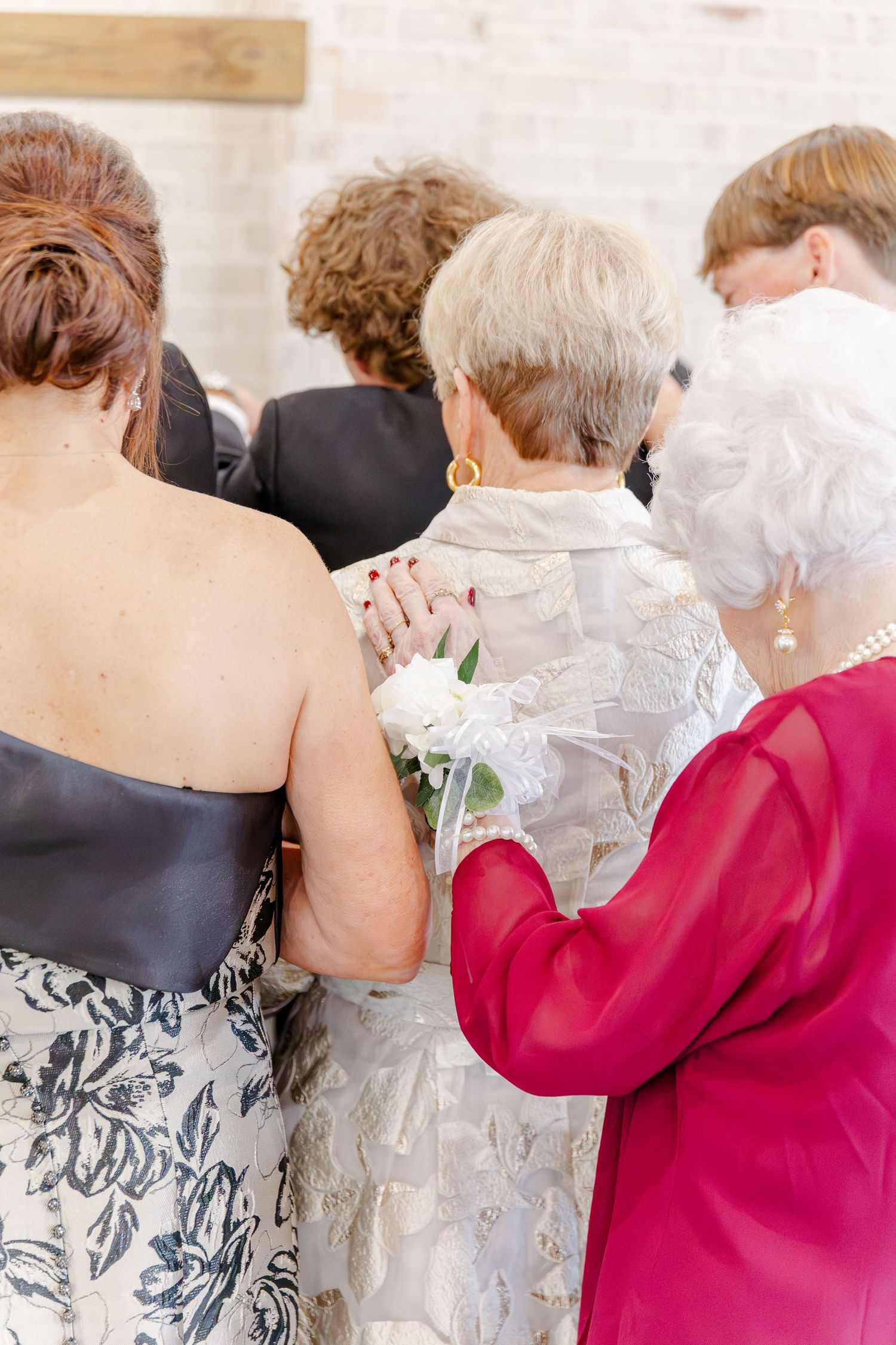 Elderly women in formal attire embrace at a wedding ceremony, one wearing a white lace dress with a floral corsage.