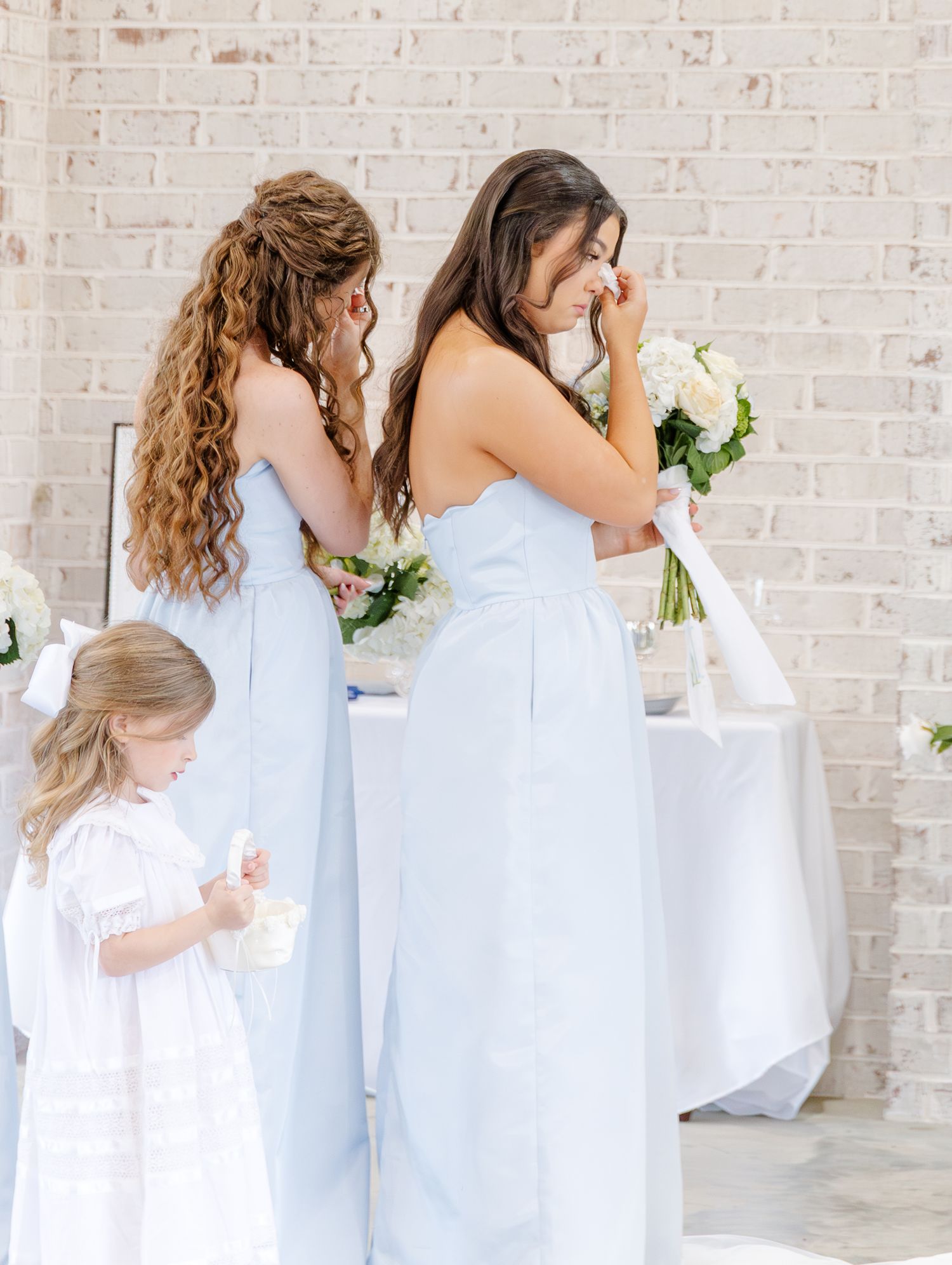 Two bridesmaids in light blue dresses and flower girl in white near floral arrangements at wedding.
