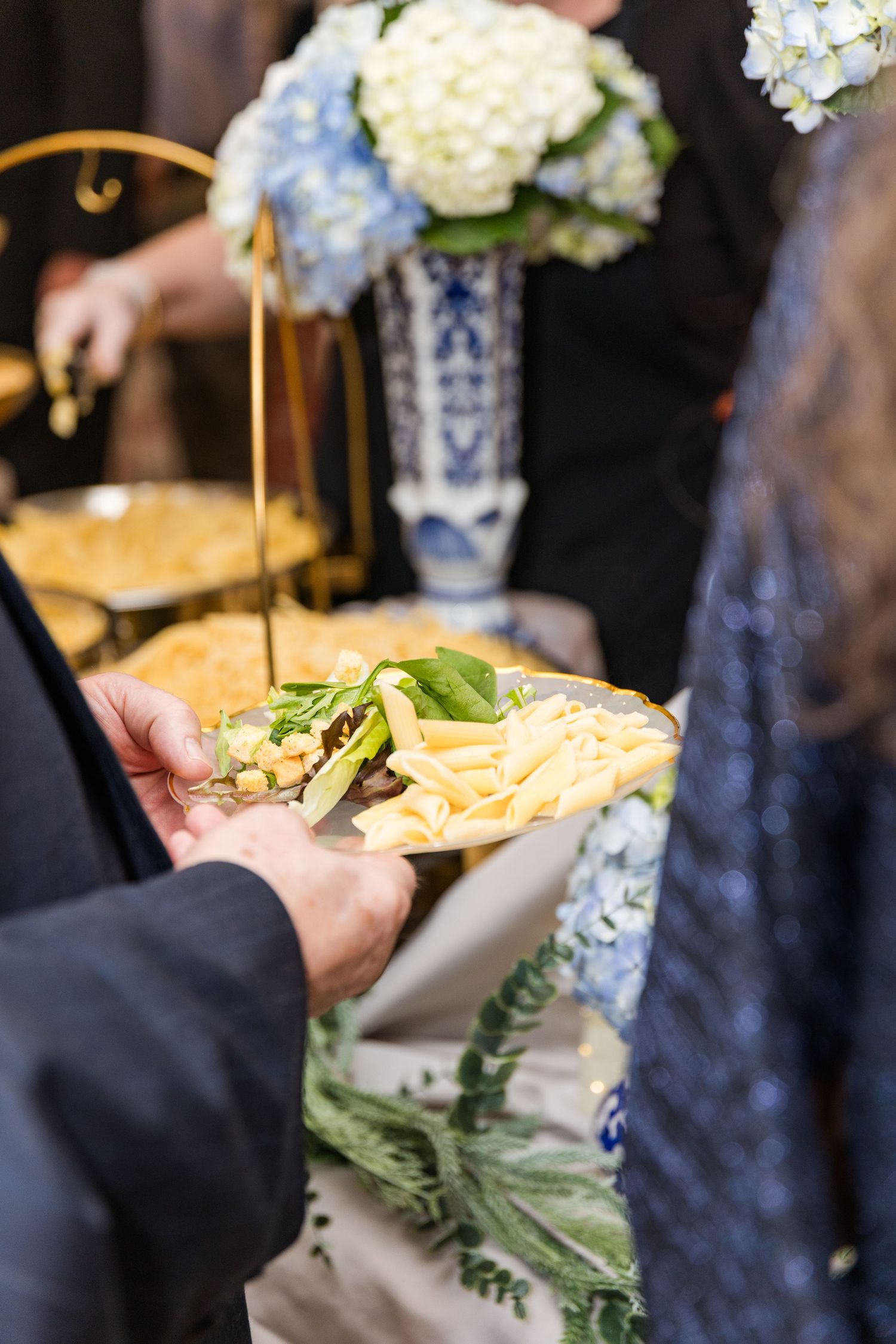 Guest holds gold plate with appetizers near blue and white vase with hydrangeas at wedding reception.