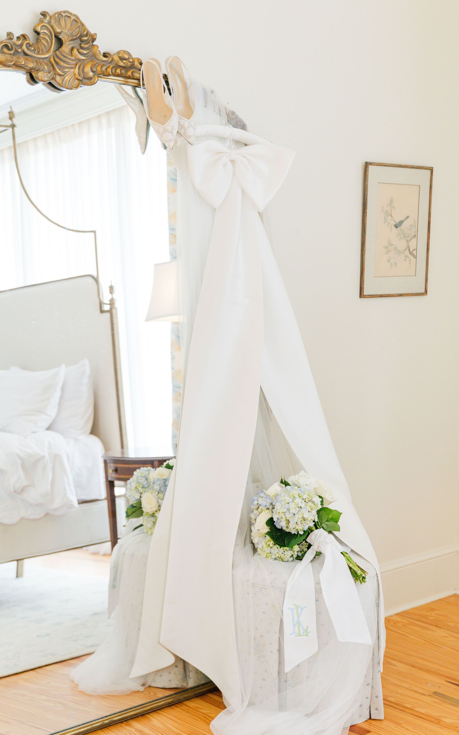 White wedding dress with large bow displayed against a gold ornate floor mirror with hydrangeas.