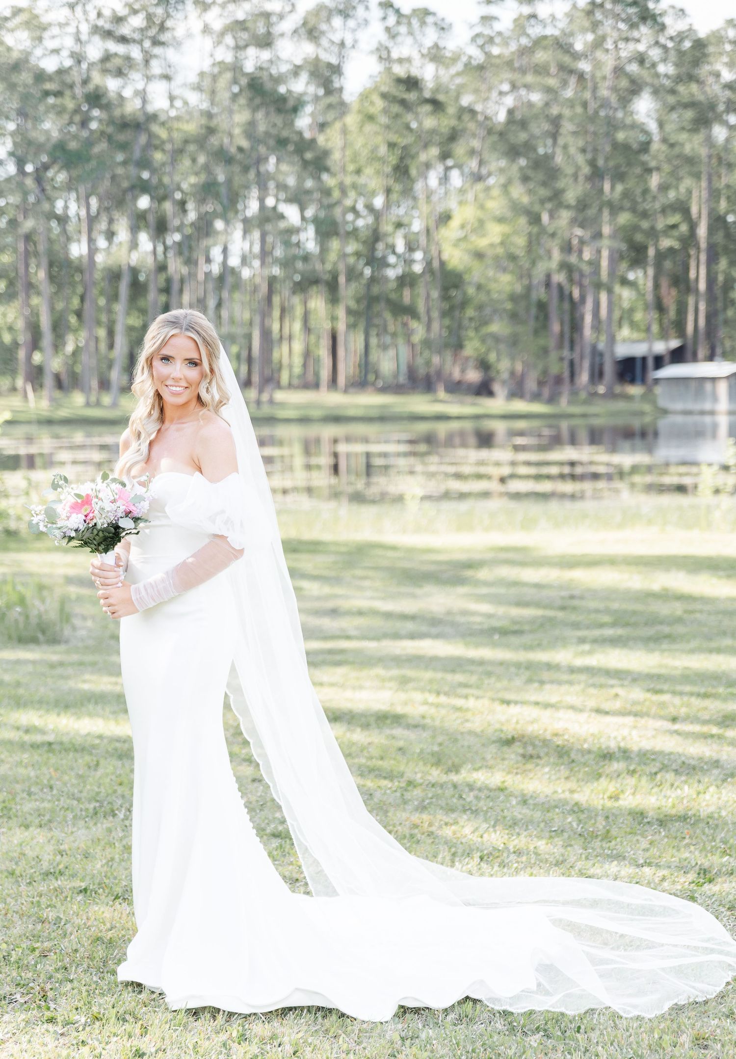 Bride in a flowing white gown with train poses in front of a lake and pine forest.