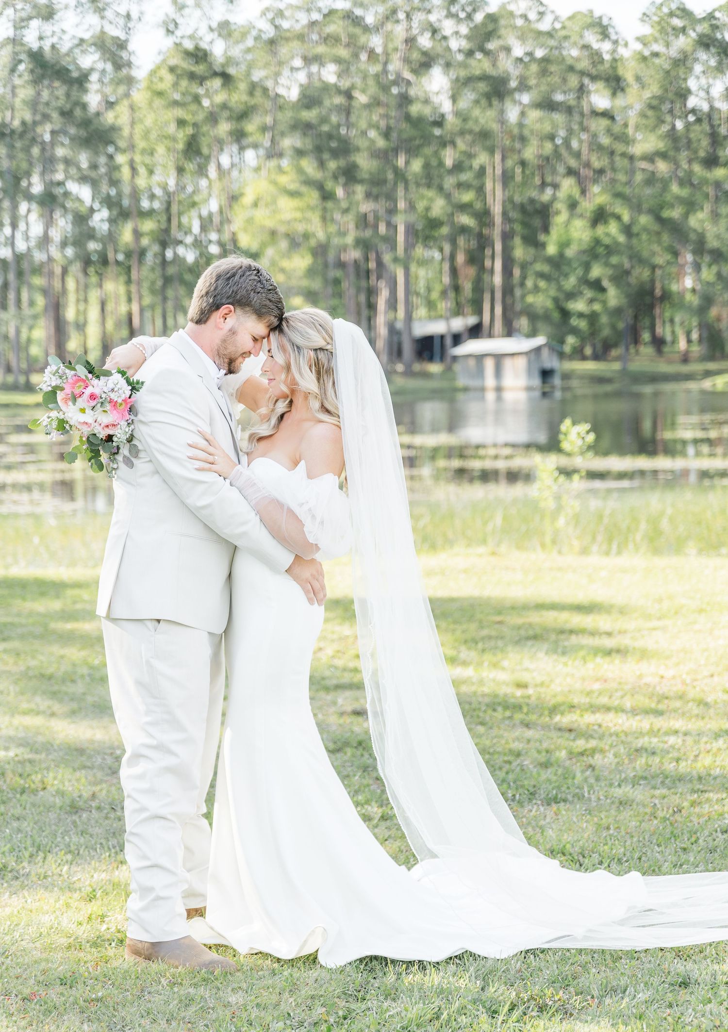 Newlyweds embrace in an outdoor wedding setting with a serene lake and pine forest backdrop.