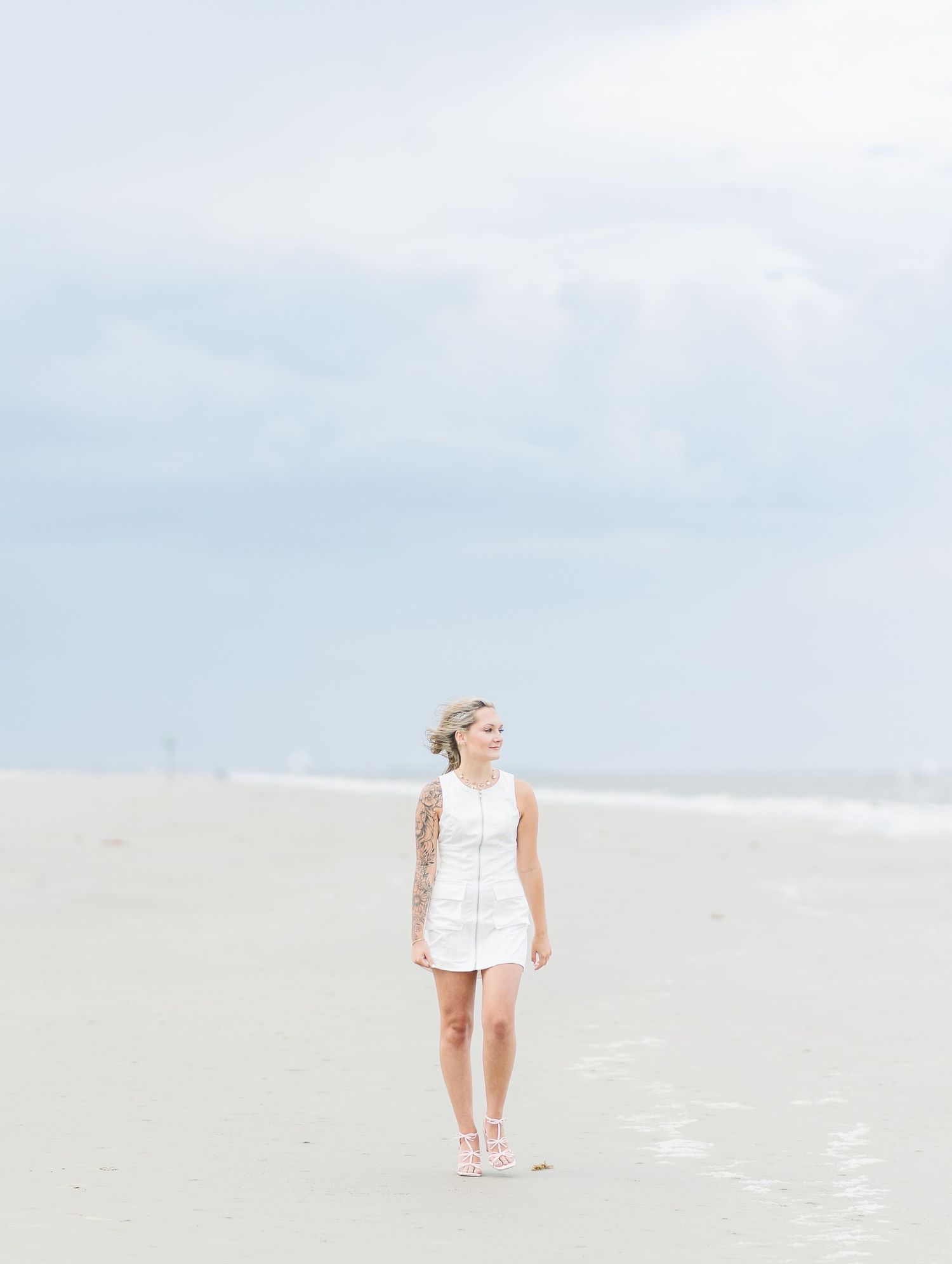 Minimalist beach scene with white dress and pink shoes creating footprints in sand.