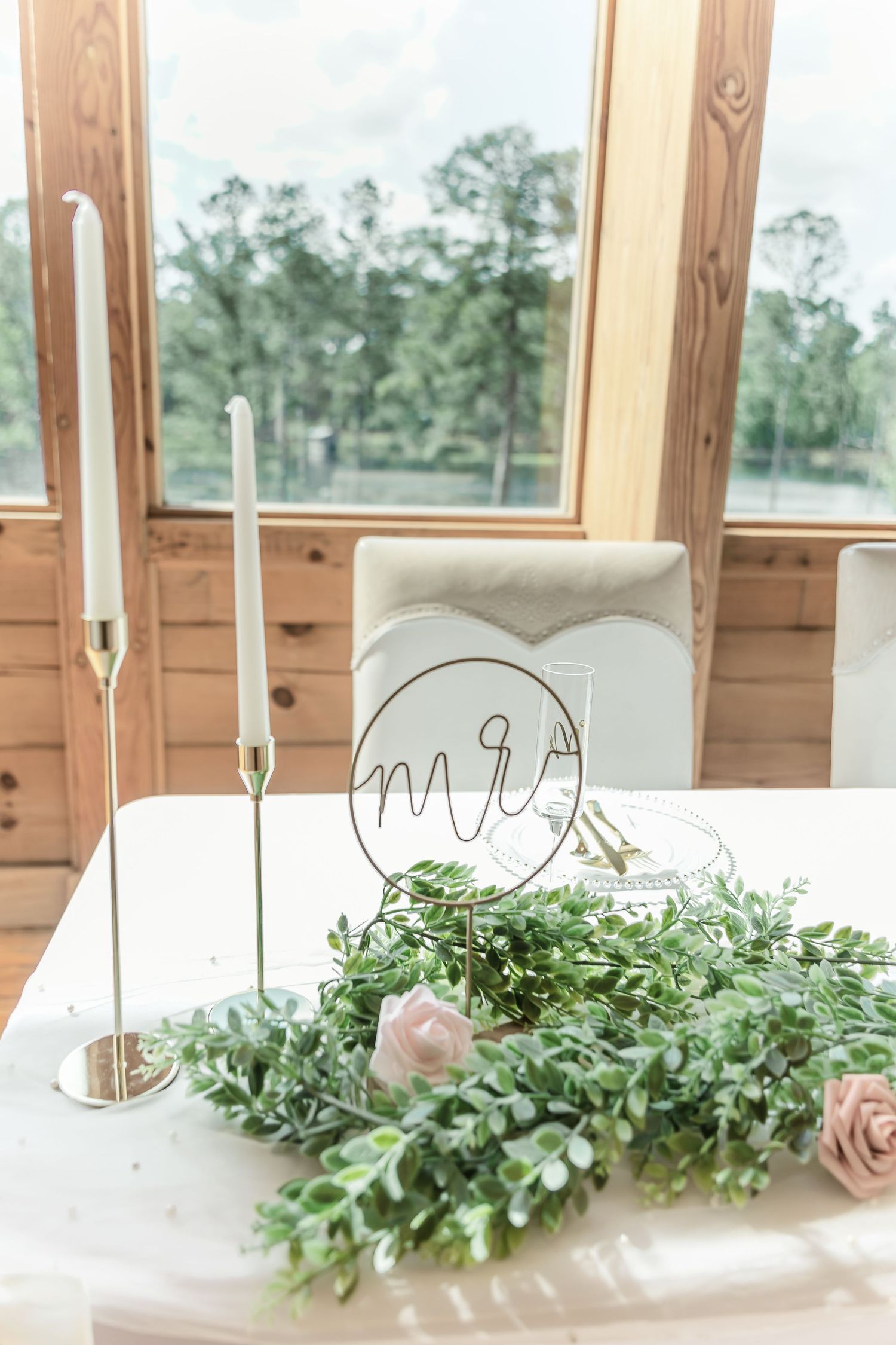 Wedding table decor with Mr & Mrs sign, white candles, and eucalyptus greenery near window.