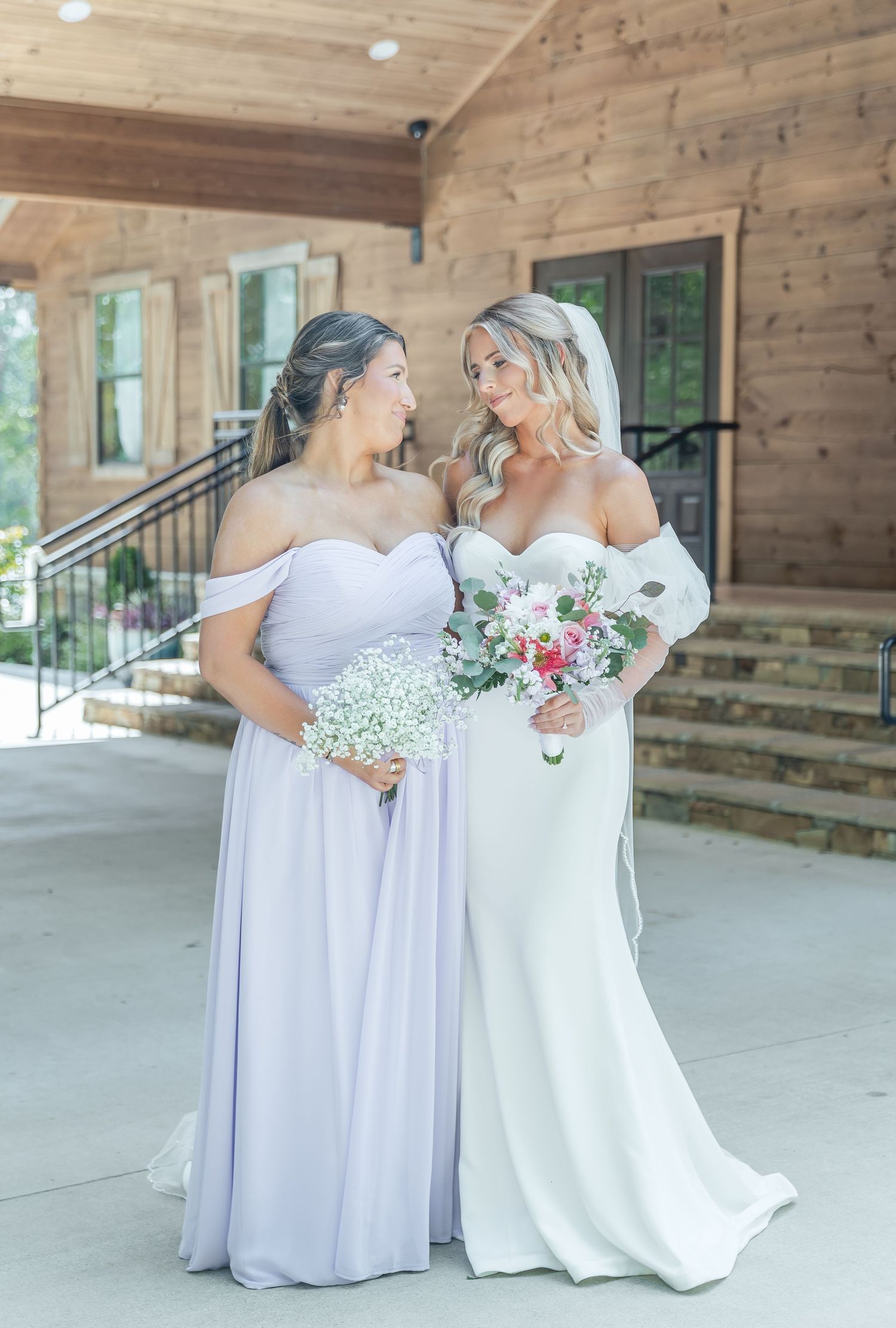 Two women share a candid moment in wedding attire holding floral bouquets on a rustic porch.