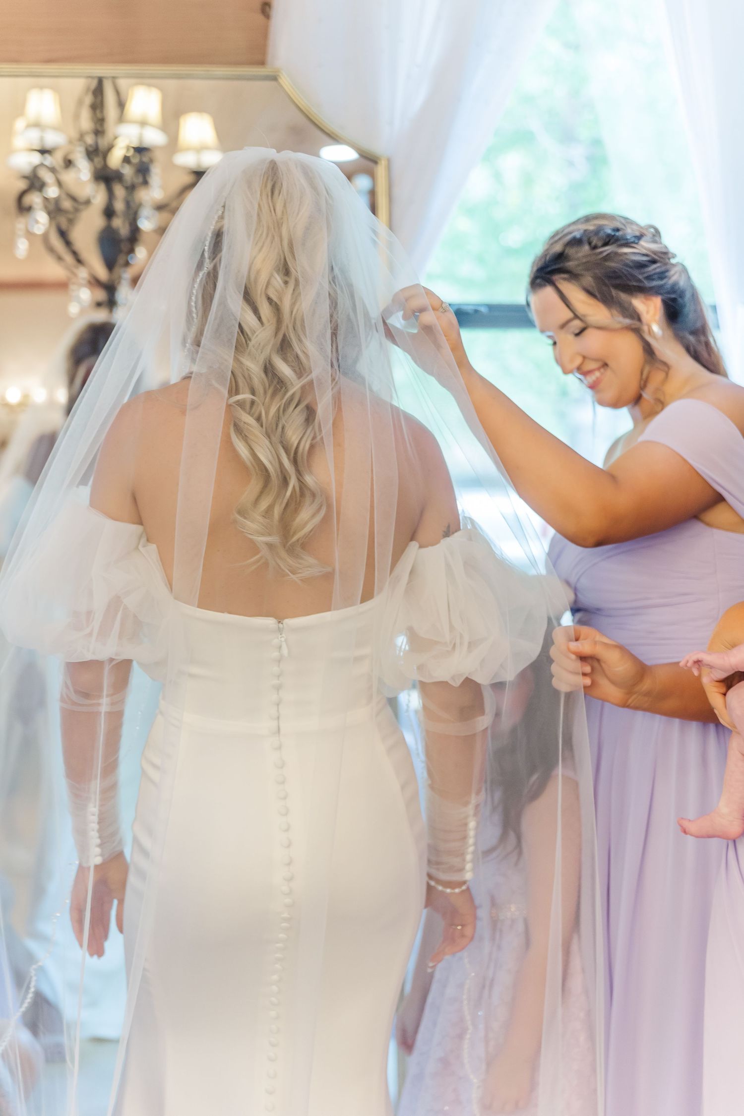 A bridesmaid in lavender helps adjust the bride's veil near a sunny window.