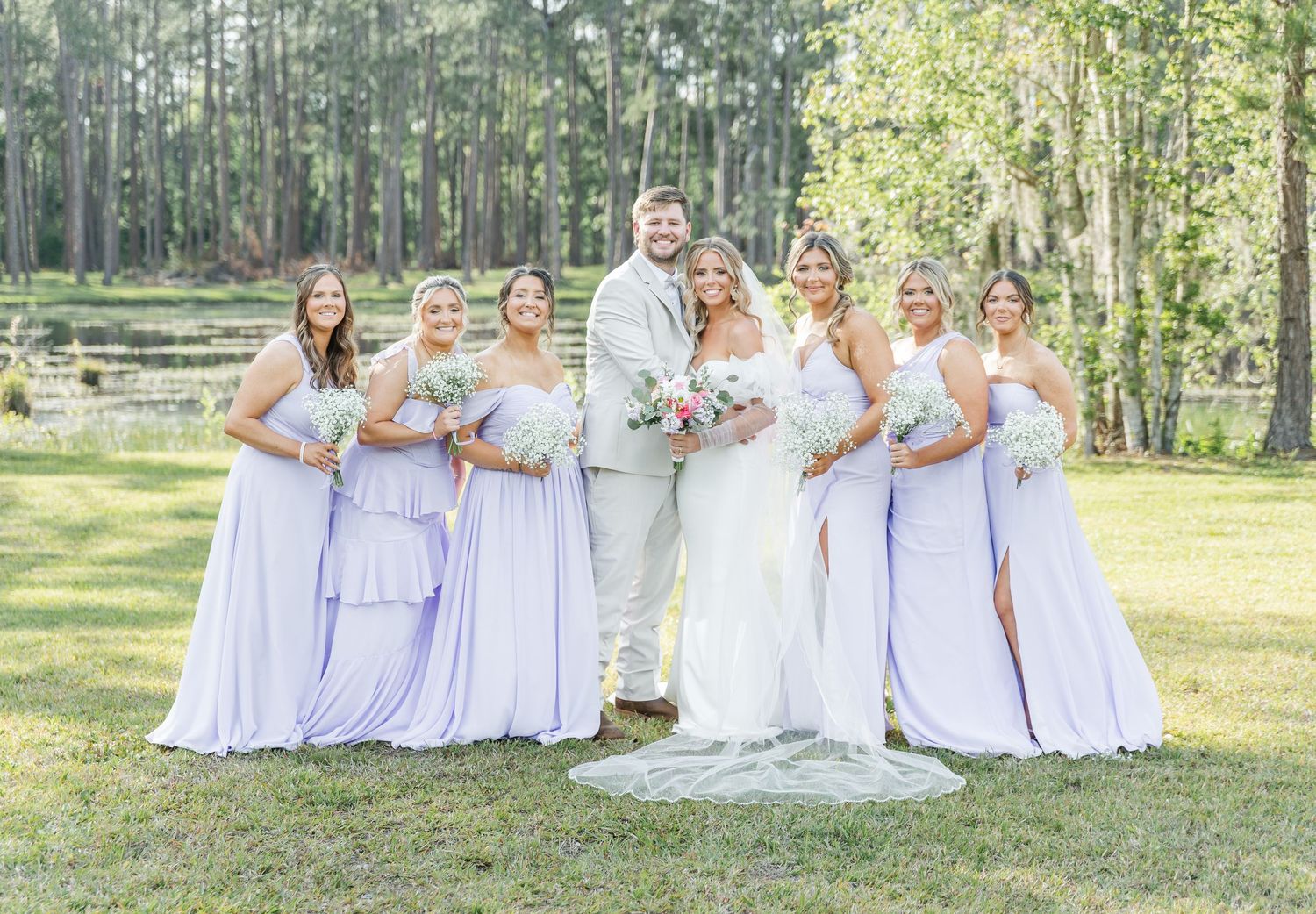 Wedding party in coordinated white and lilac attire poses on sunny lawn.
