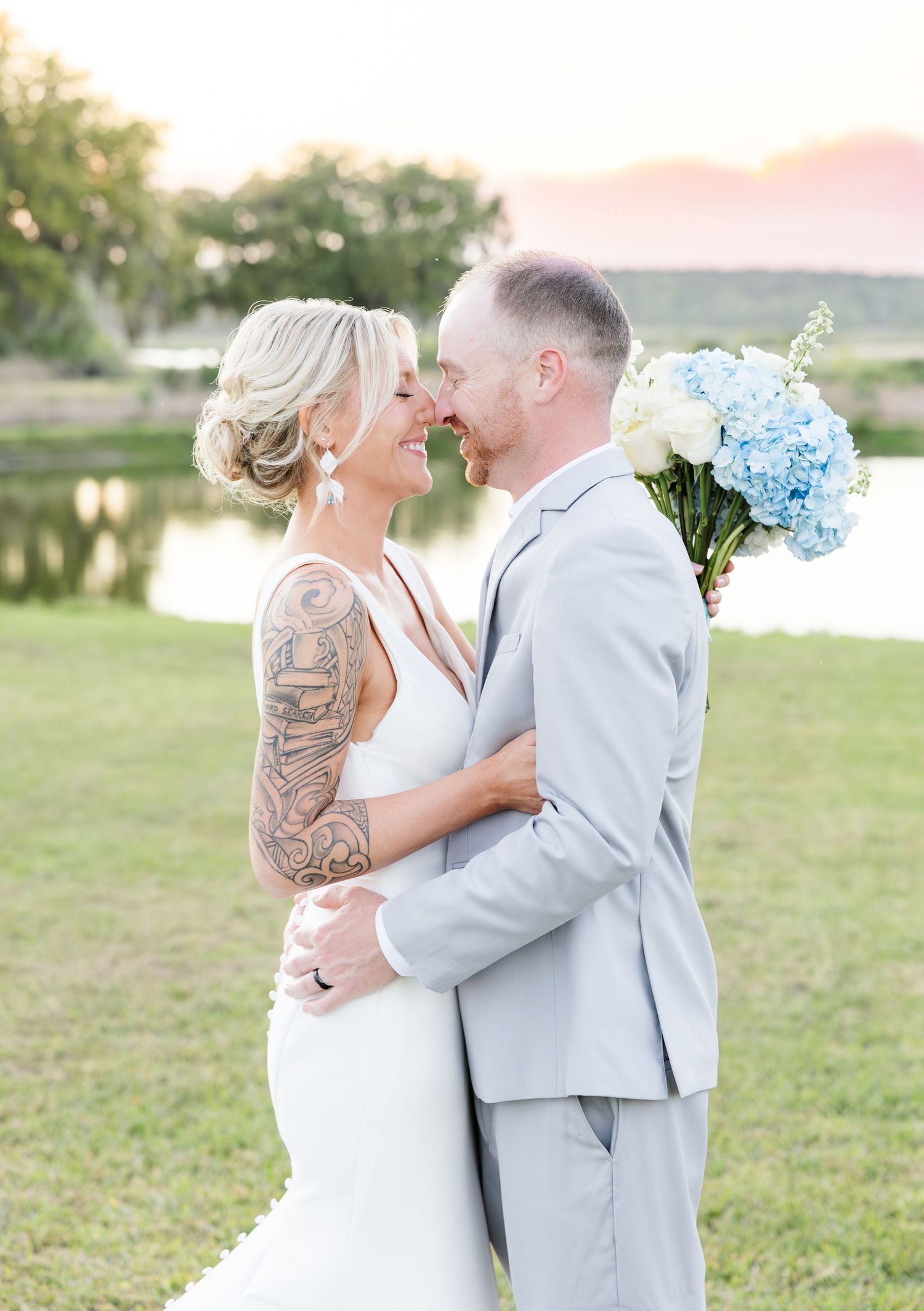 Bride with tattoo sleeve and groom in gray suit share romantic moment holding blue hydrangea bouquet at outdoor wedding.