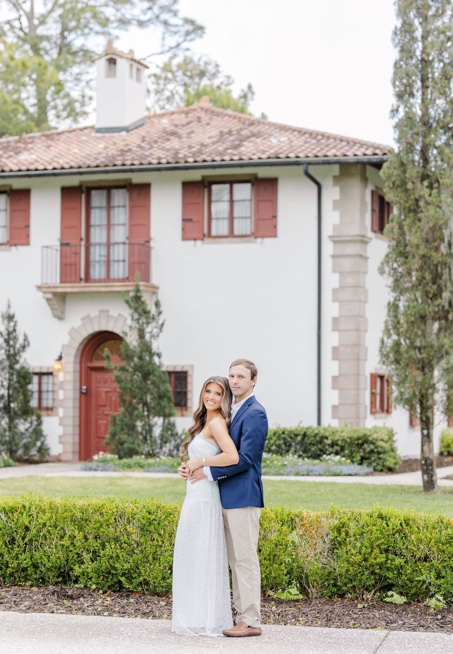 Couple embracing in front of white Mediterranean-style villa with red shutters and tile roof.