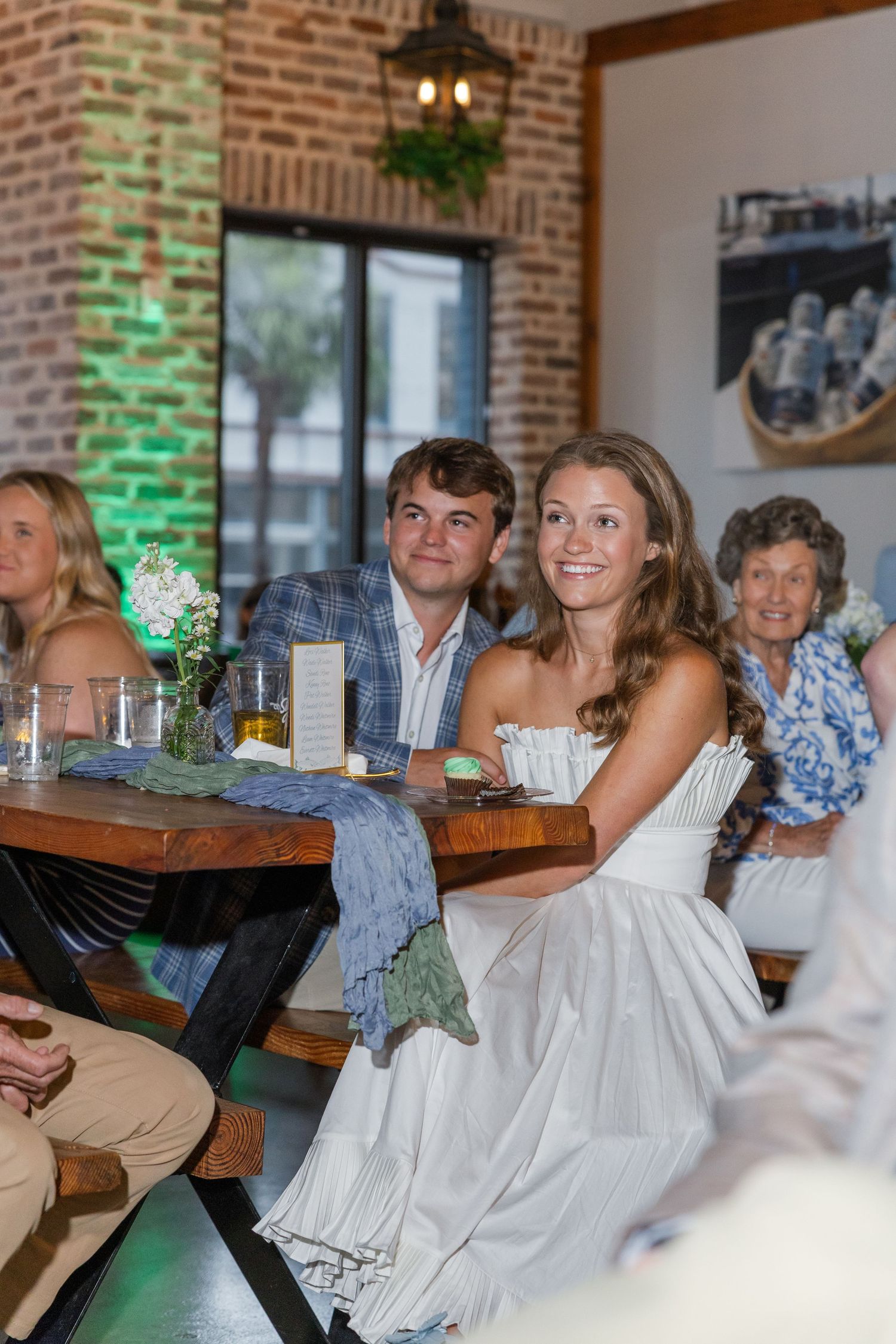 Smiling bride in white dress sits with groom at rustic wooden table during wedding reception in brick-walled venue.