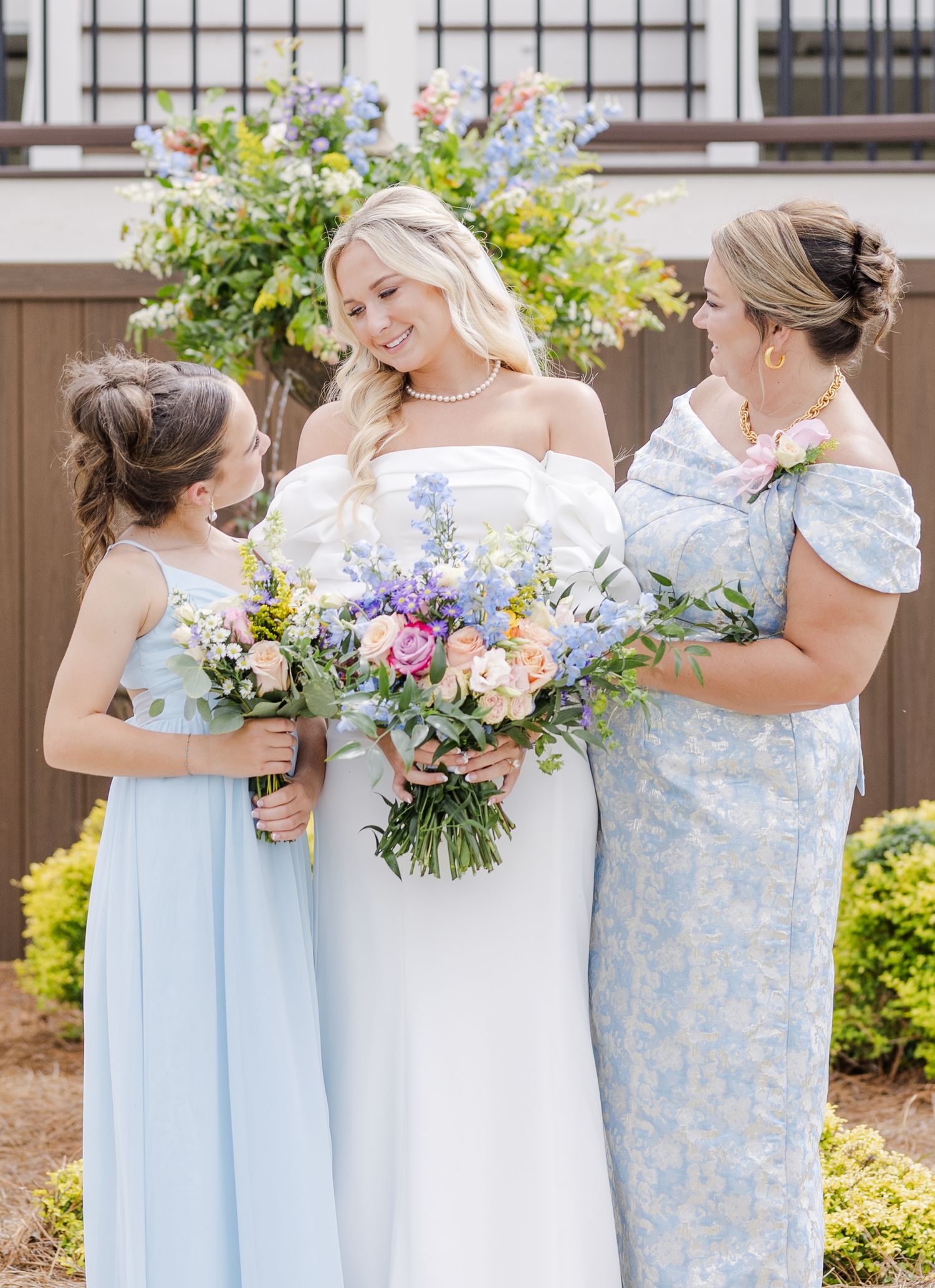 Bride in white off-shoulder gown with pearl necklace holding colorful bouquet flanked by family.