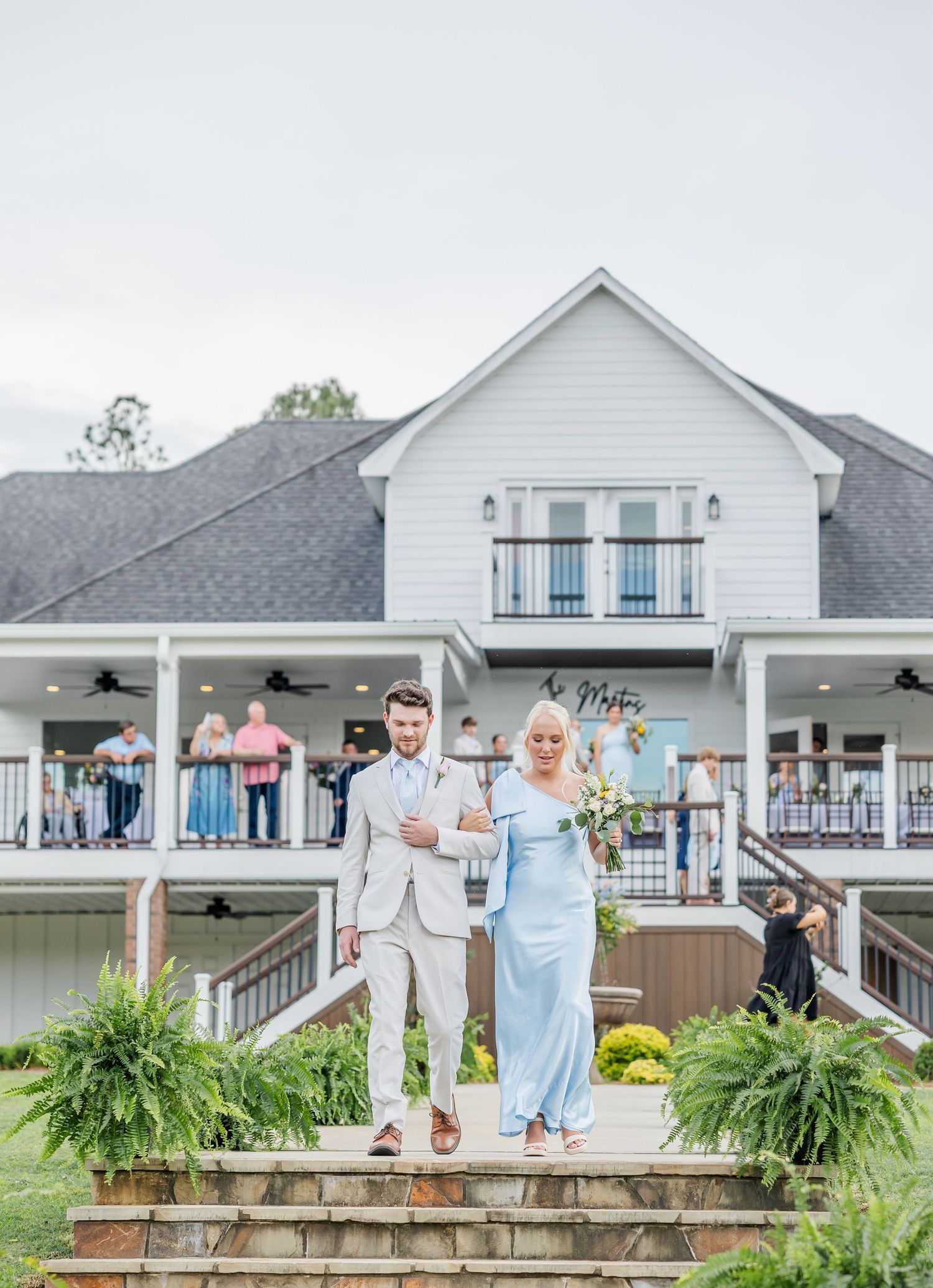 Groomsman escorting woman in light blue gown down steps outside white wedding venue.