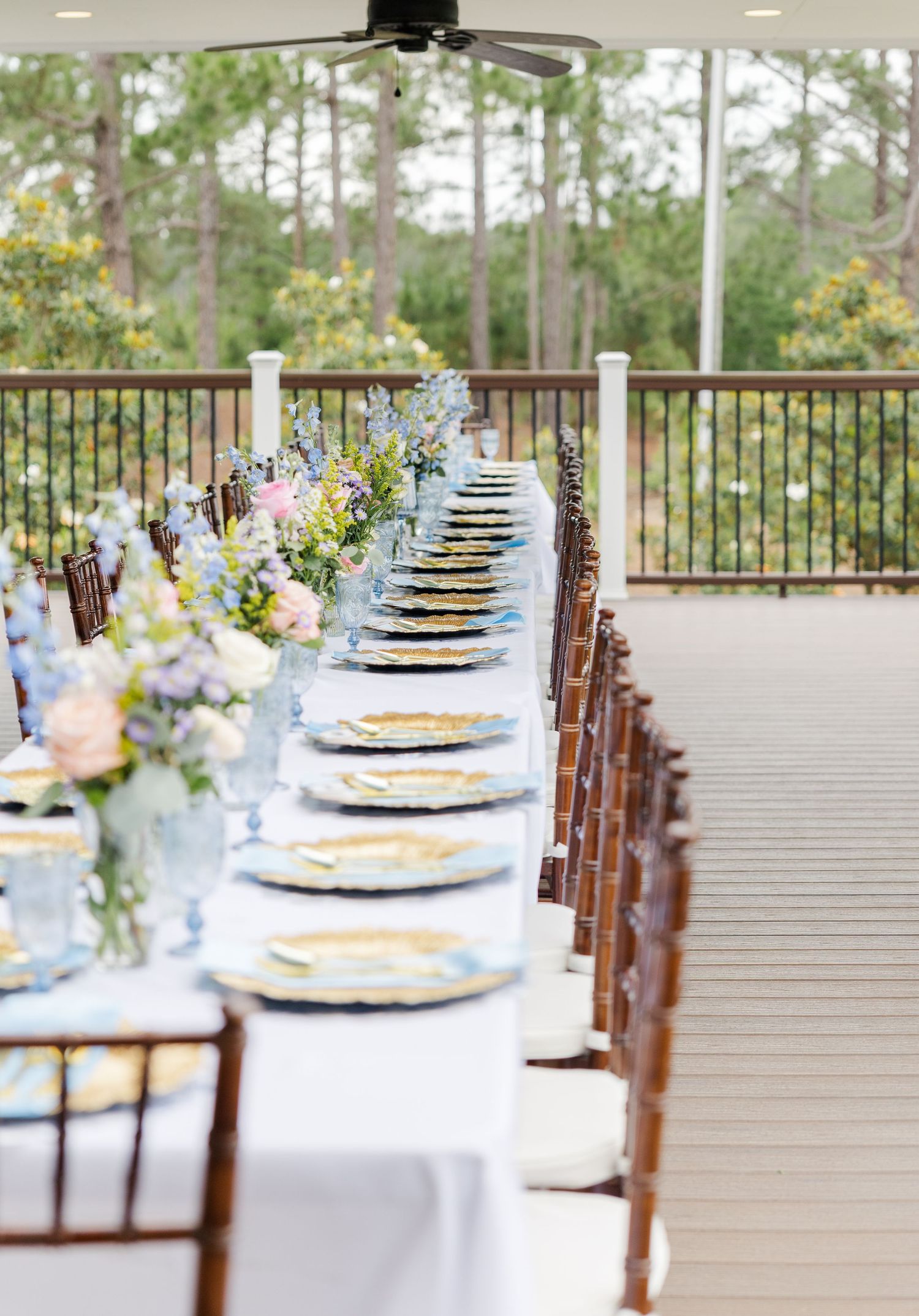 Elegant outdoor wedding reception table set with blue chargers and colorful floral centerpieces on a deck.