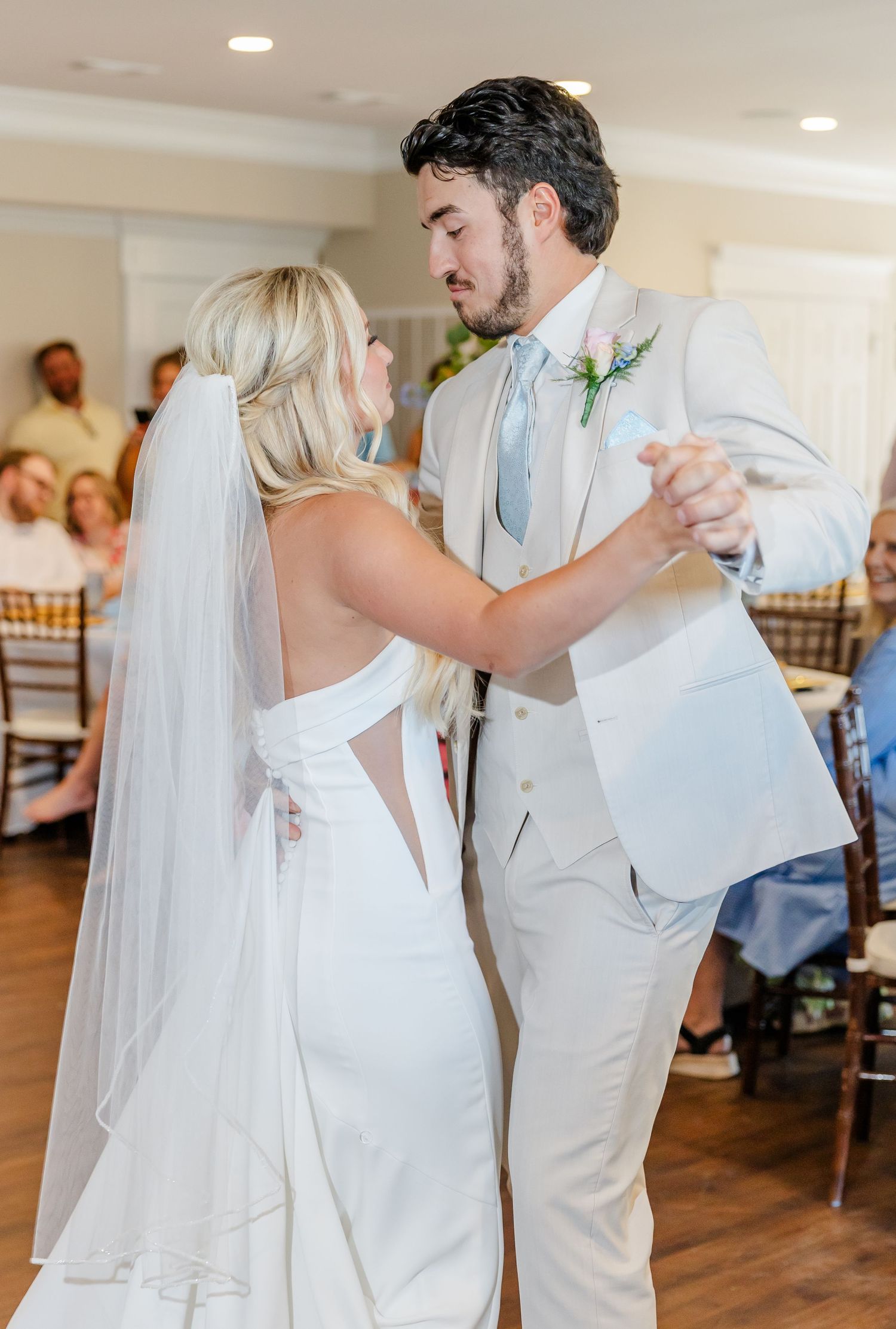 Bride and groom sharing first dance, bride in white gown with veil, groom in tan suit.