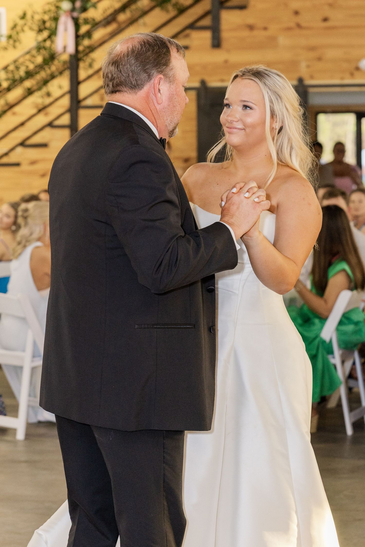 Smiling bride in strapless white gown holds hands with father during wedding reception dance.