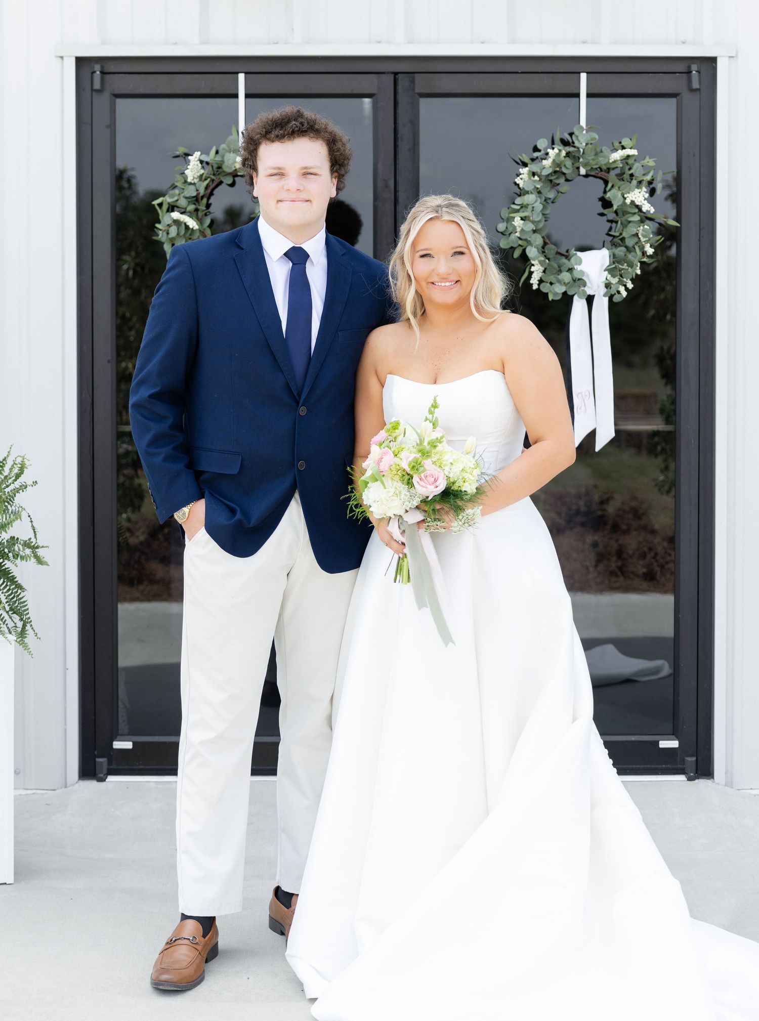Bride in strapless white gown holding bouquet poses with groomsman in navy suit at wedding venue.