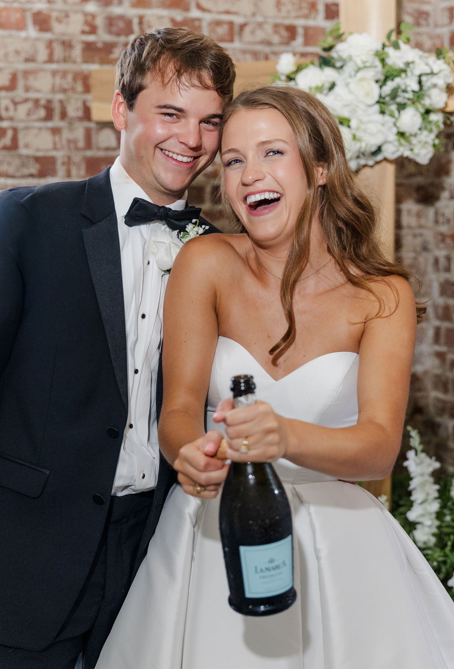 Laughing newlyweds celebrate with champagne bottle, bride in strapless gown and groom in tuxedo.