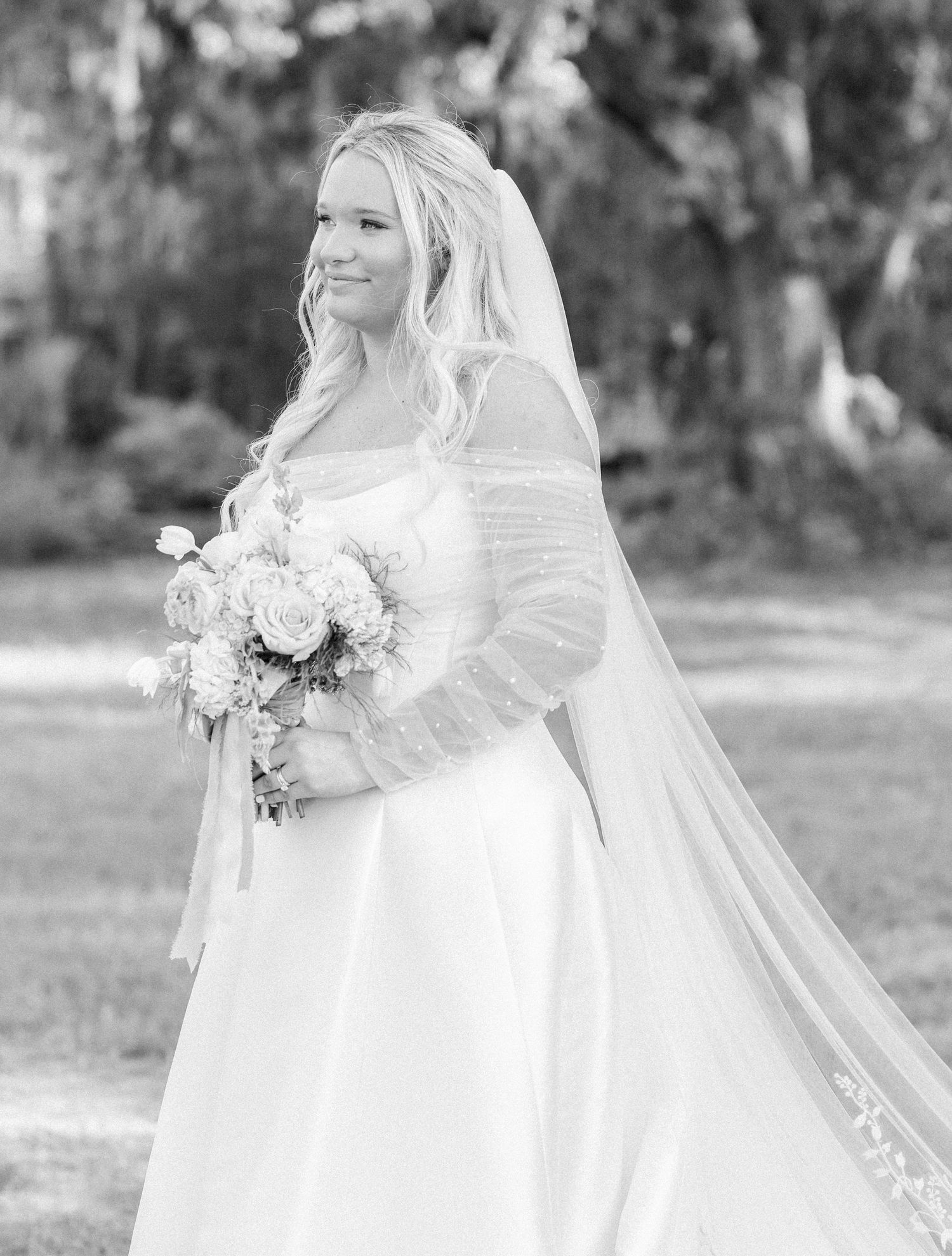 Black and white portrait of bride in wedding gown holding bouquet with flowing veil outdoors.