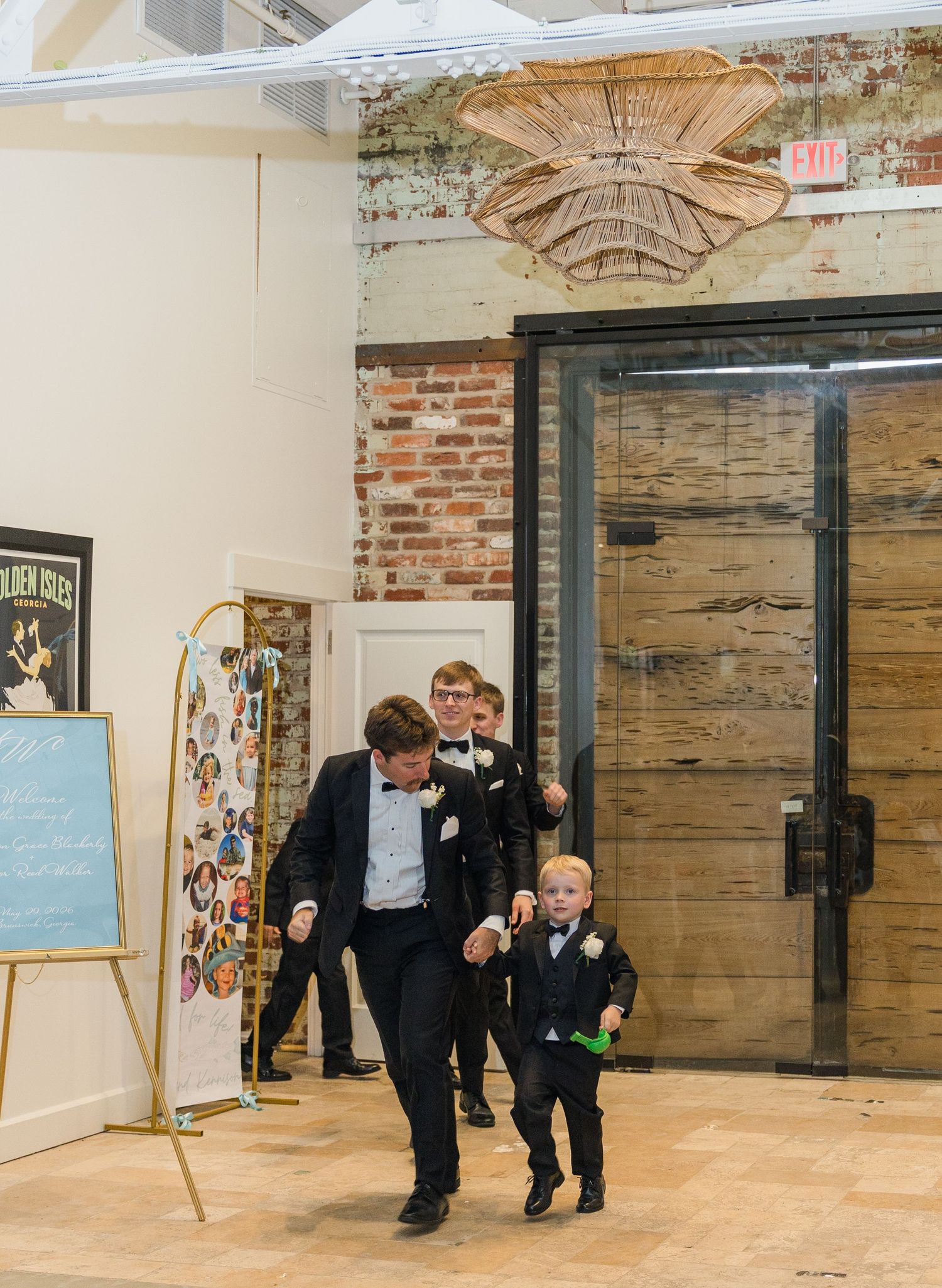 Groomsman leads excited young ring bearer through industrial chic wedding venue entrance.