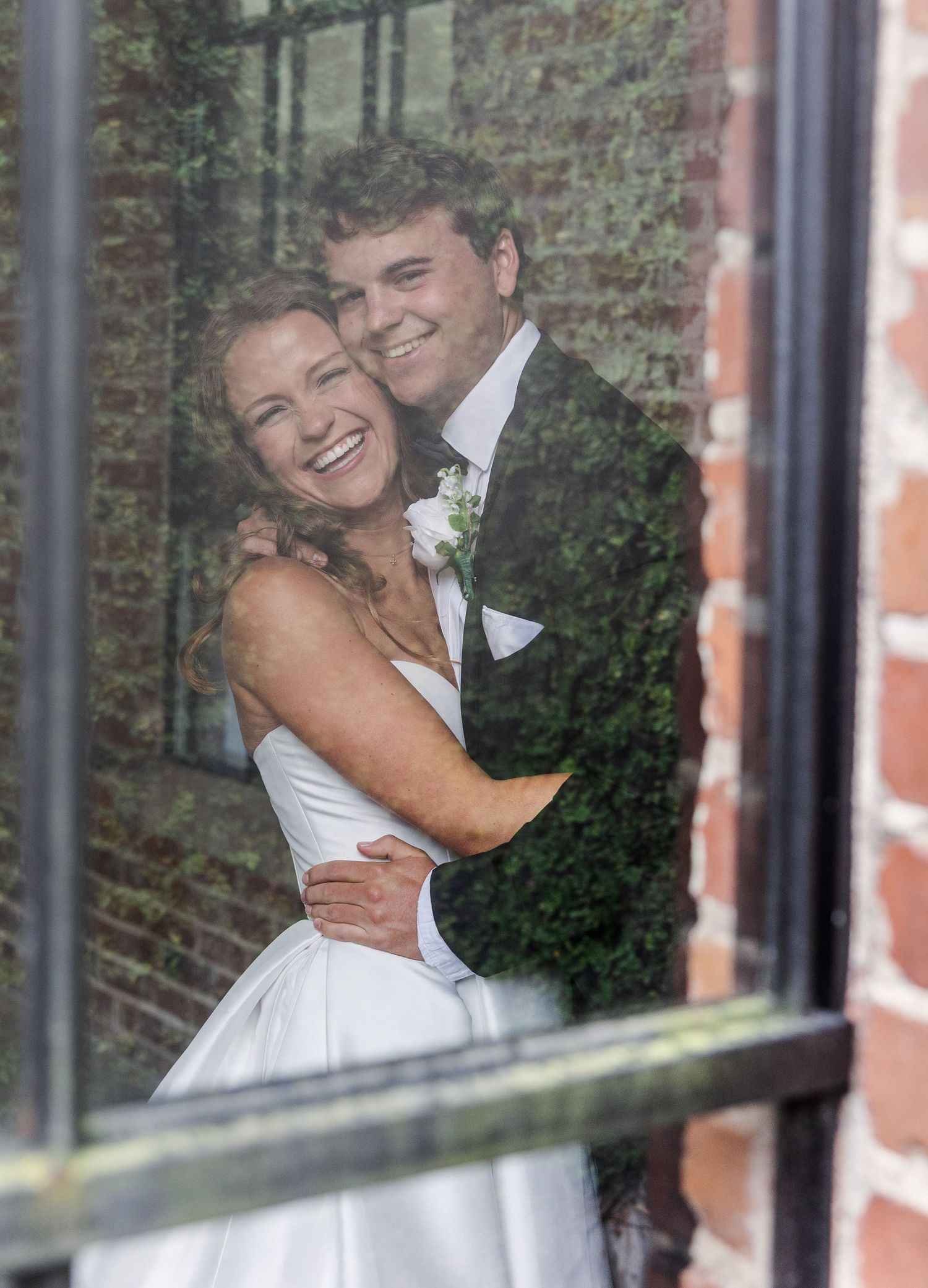 Bride and groom smiling and embracing by a brick wall window on their wedding day.