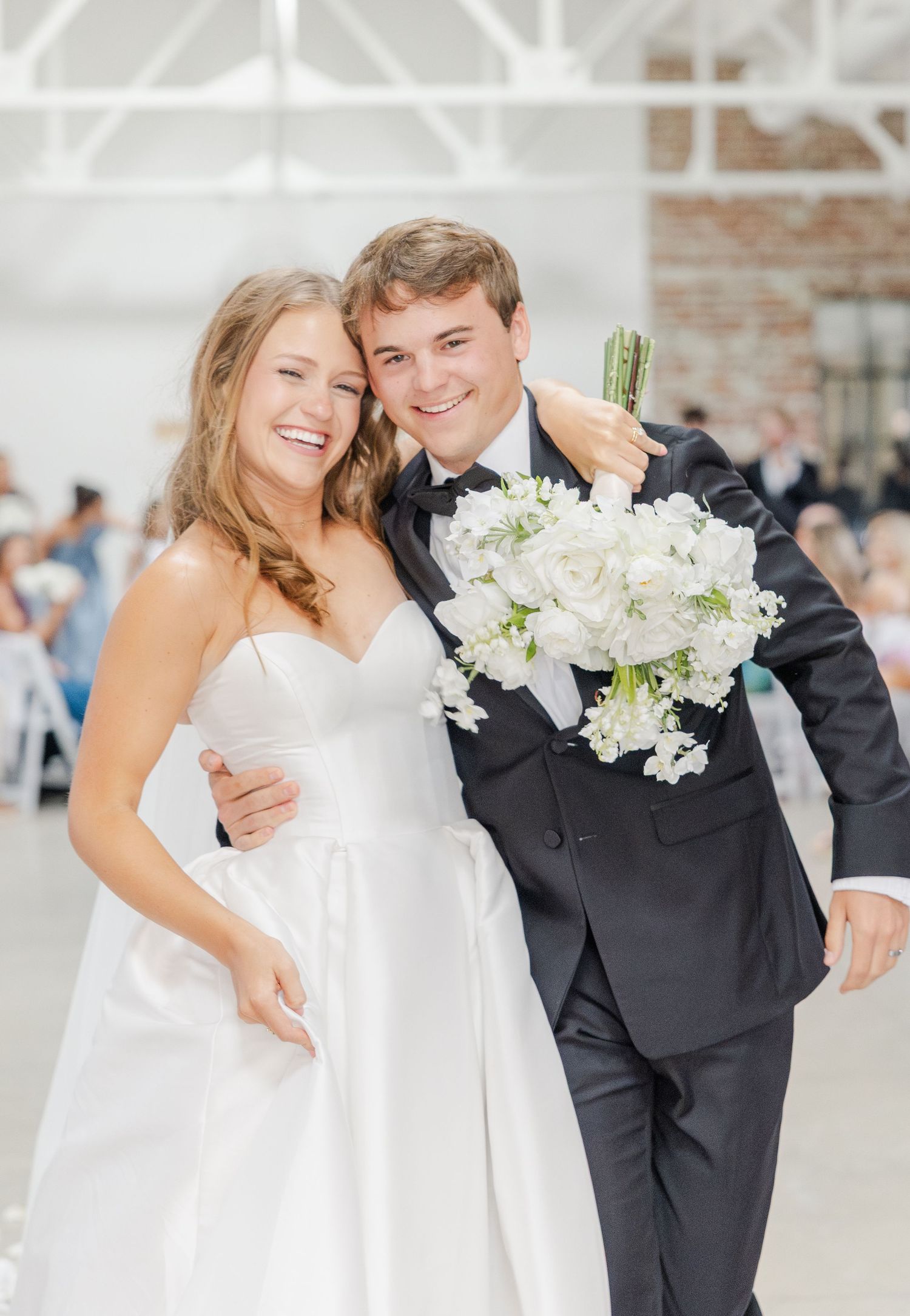 Smiling bride in strapless white gown and groom in black tuxedo holding white bouquet after wedding ceremony.