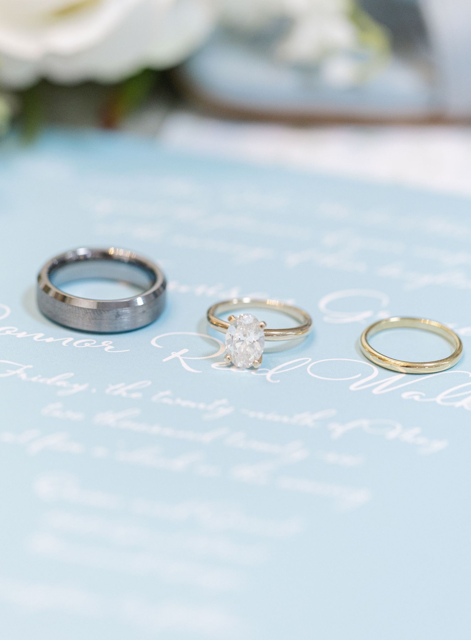 Wedding rings displayed on blue invitation: tungsten band, diamond solitaire, and gold band.