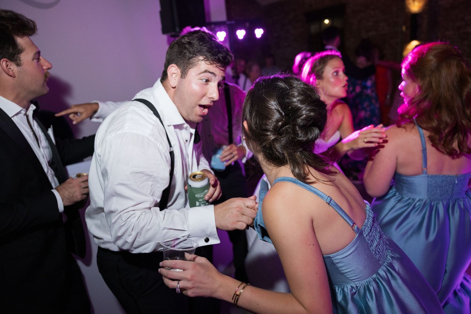 Surprised wedding guest in white shirt with suspenders reacting expressively on colorful reception dance floor.