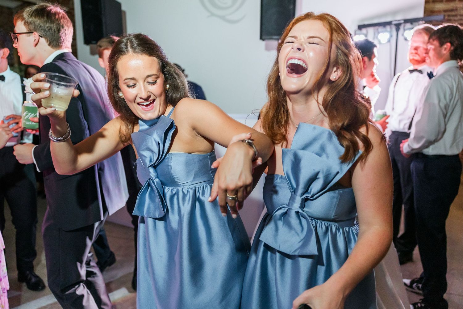 Two bridesmaids in blue bow dresses laughing and dancing joyfully at wedding reception celebration.