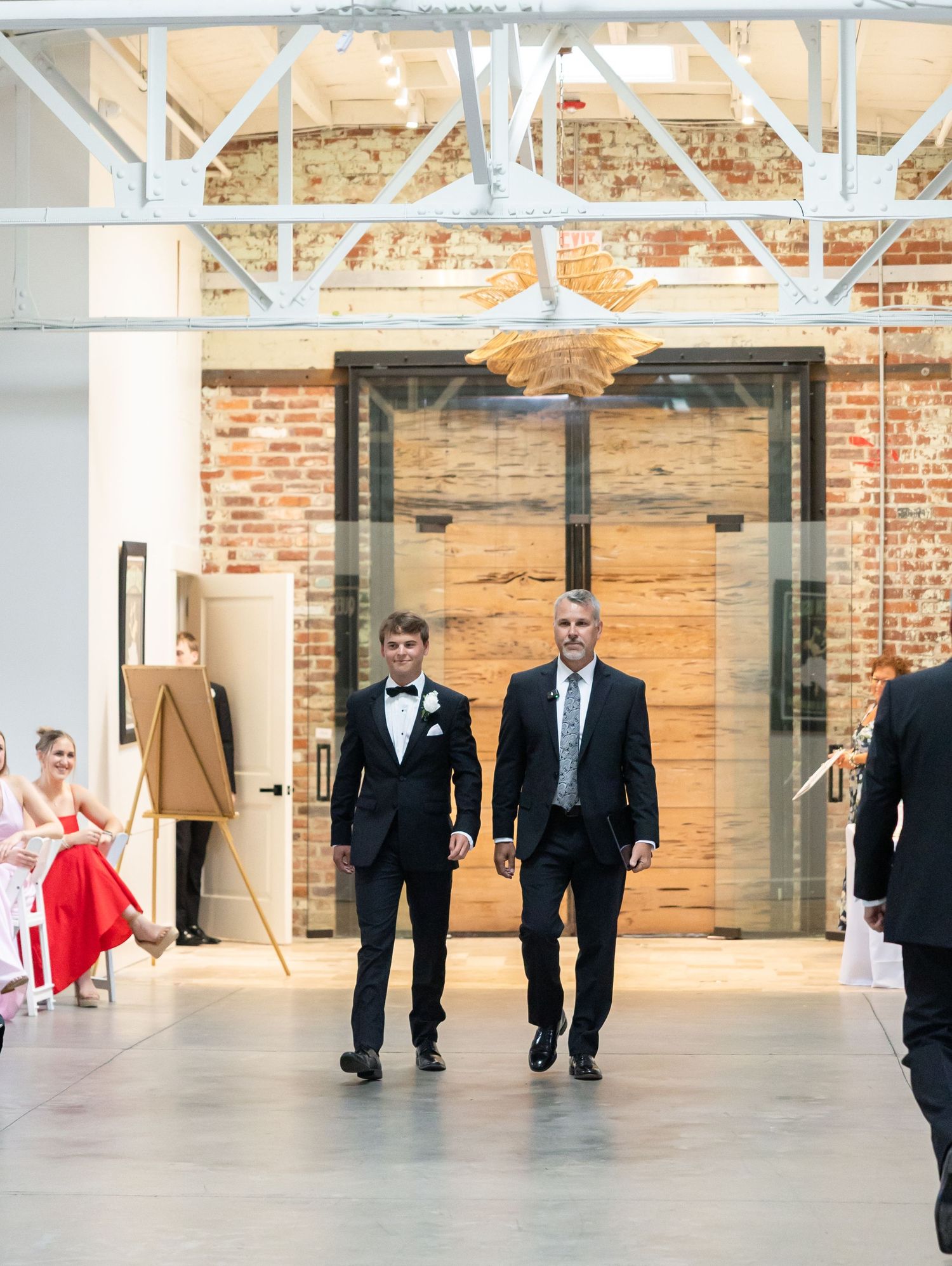 Groom and father walking down aisle in industrial wedding venue with exposed brick and steel beams.