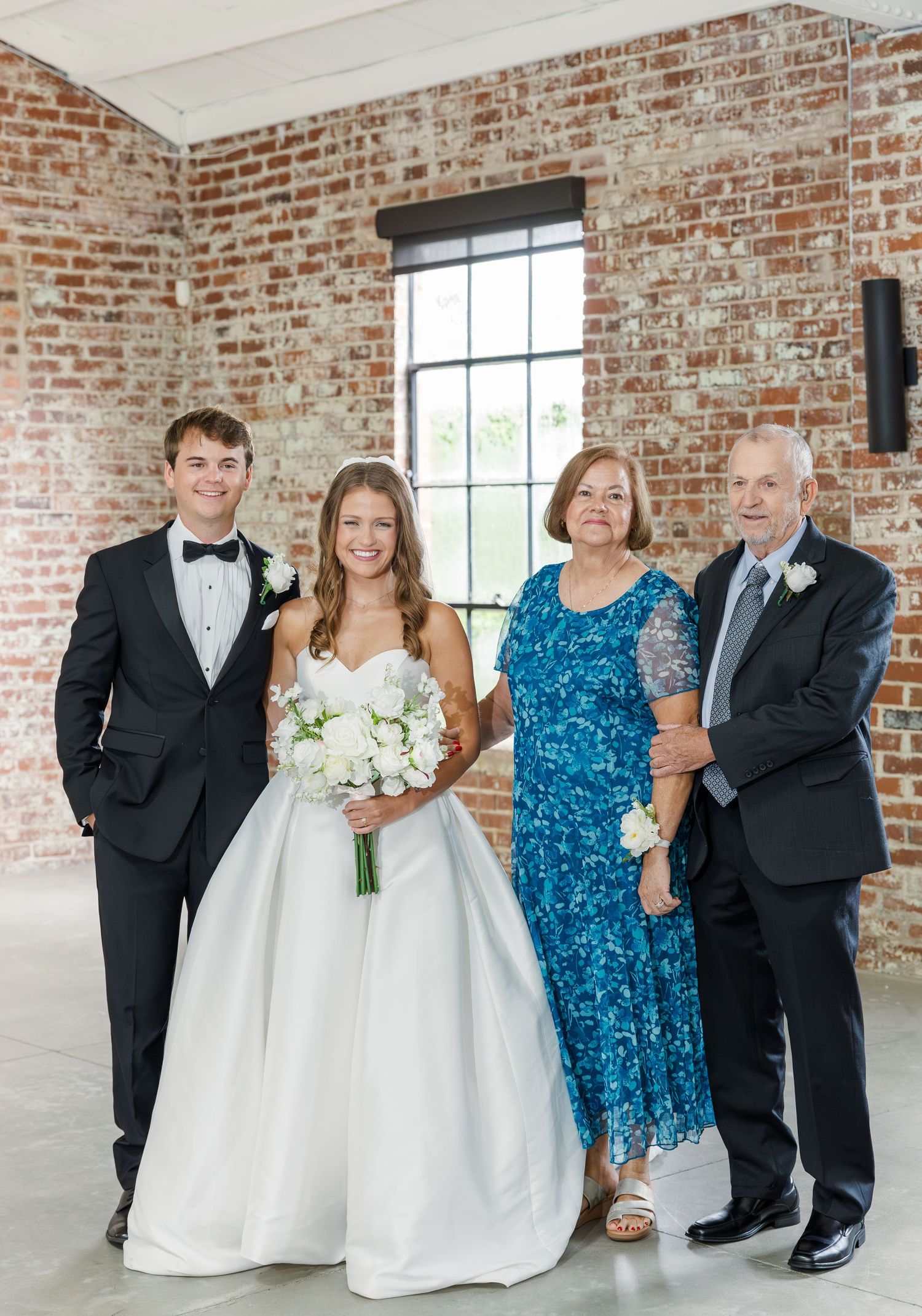 Bride and groom posing with grandparents in elegant wedding attire in brick venue.