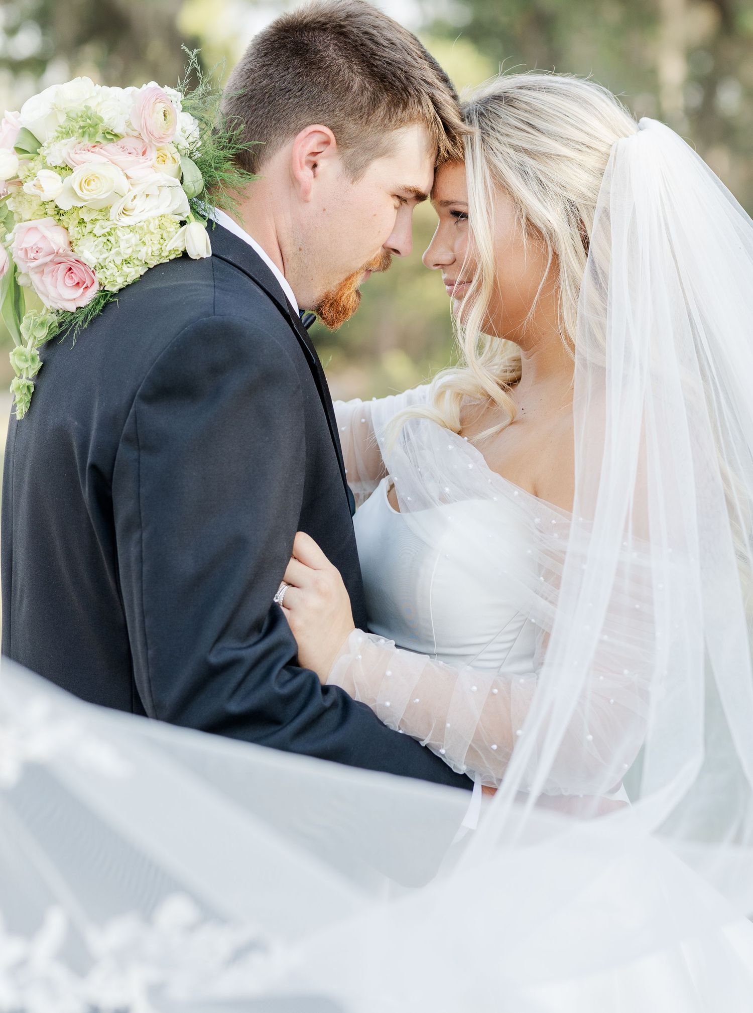 Bride and groom sharing an intimate moment, with her lace veil flowing and a pink floral bouquet visible.