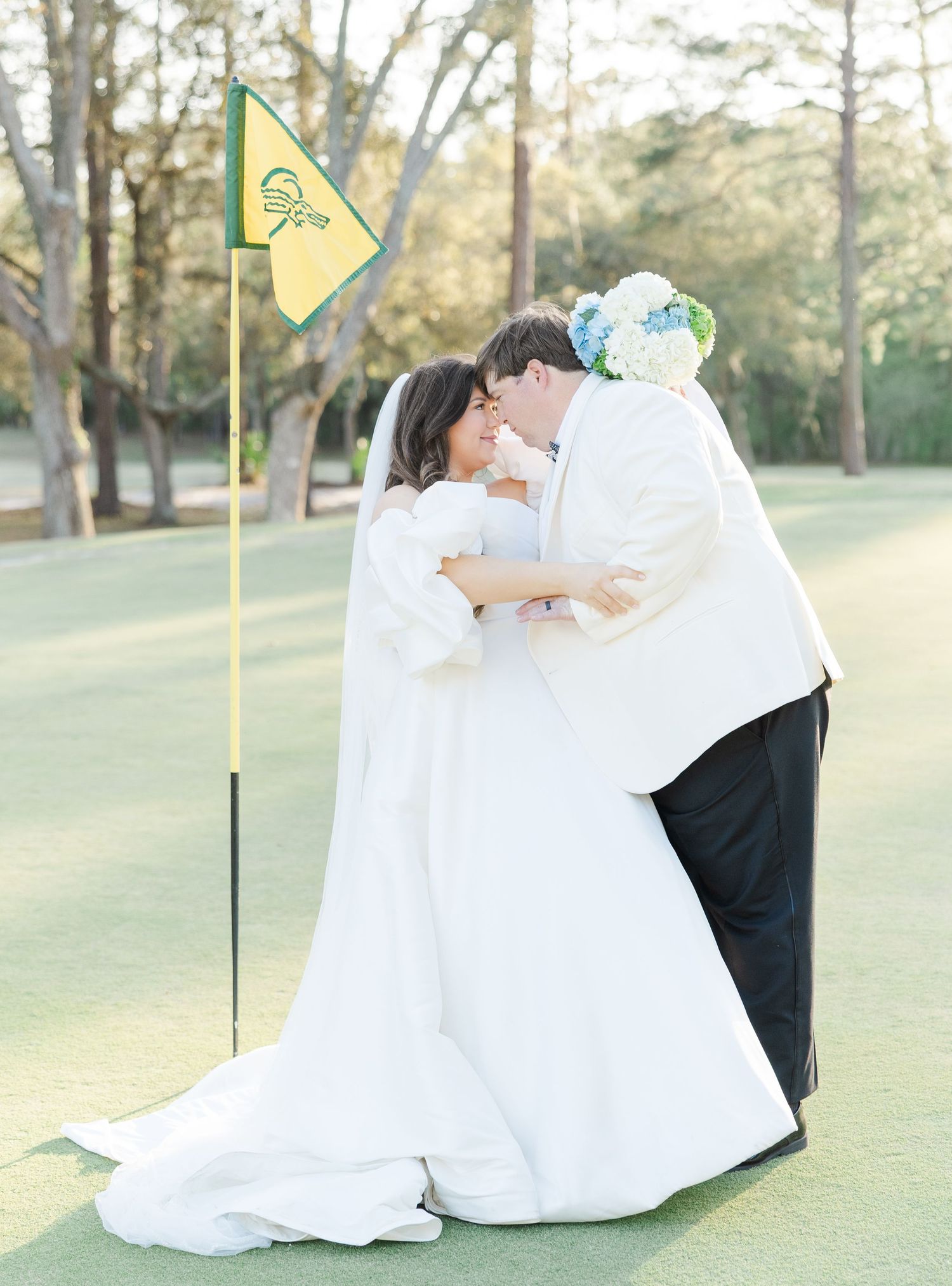 Bride and groom share a romantic kiss on a golf course green beside a yellow flag on their wedding day.