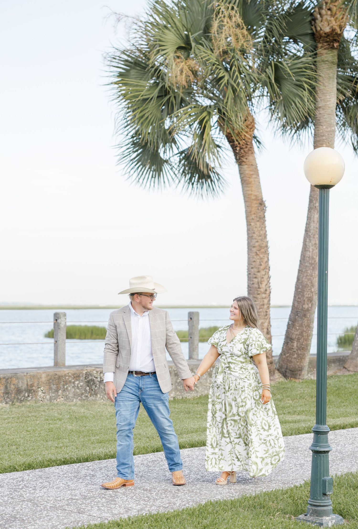 Couple holding hands walking near palm trees and waterfront, man in cowboy hat and blazer, woman in floral dress.