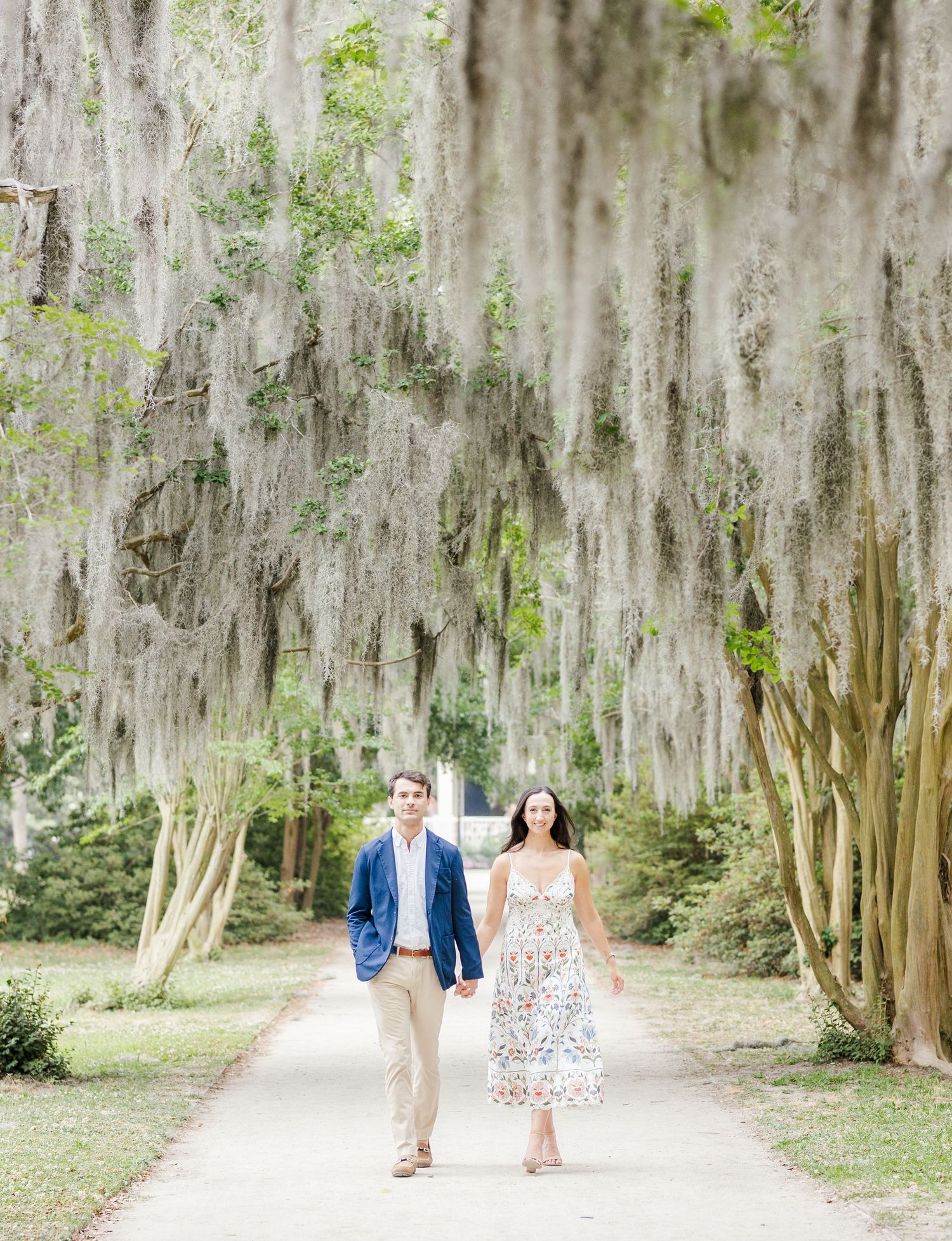 Couple walks hand-in-hand down a moss-draped oak alley, woman in floral dress, man in blue blazer.
