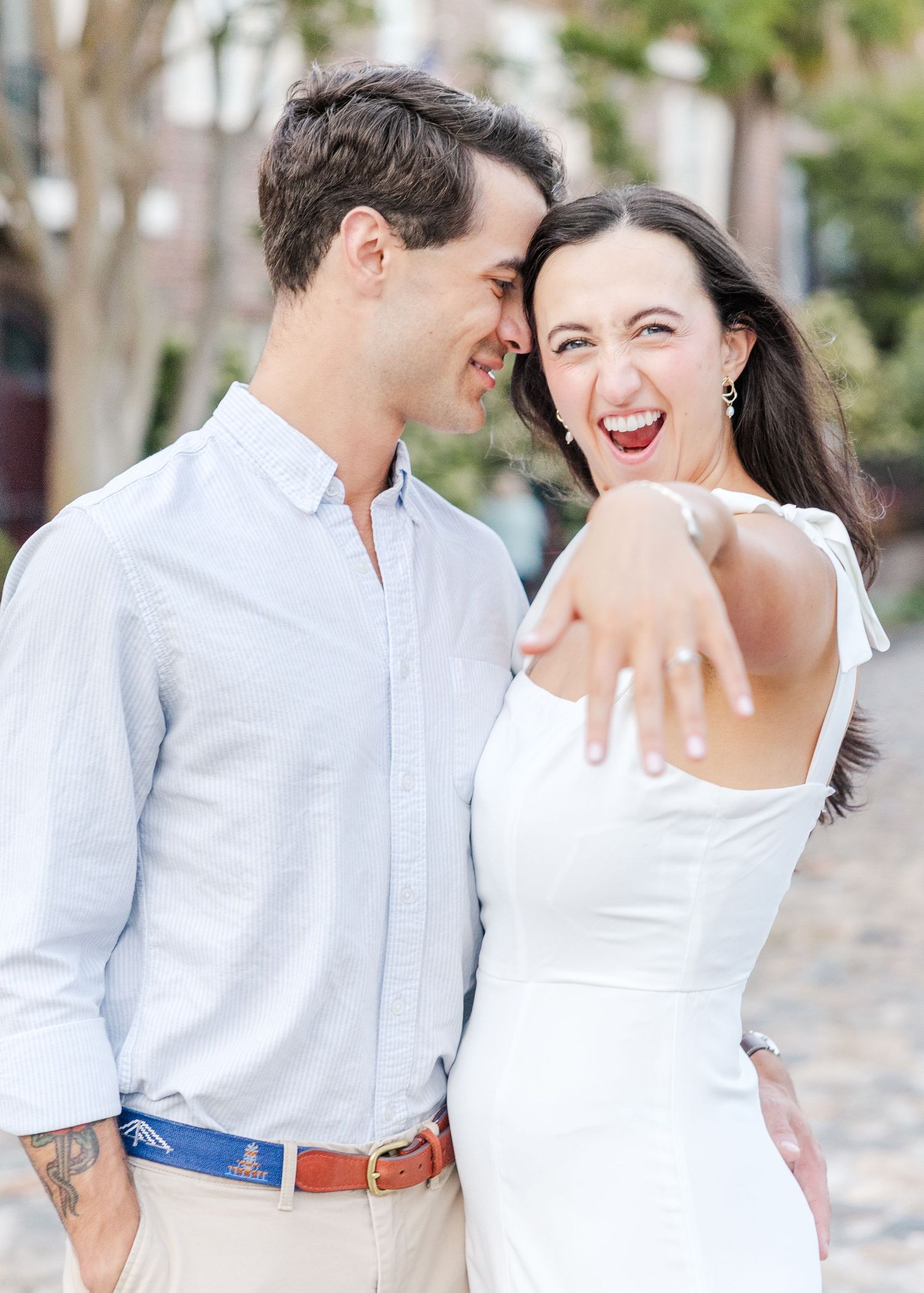 Joyful woman shows off engagement ring while laughing with her fiance on a cobblestone street in Charleston.
