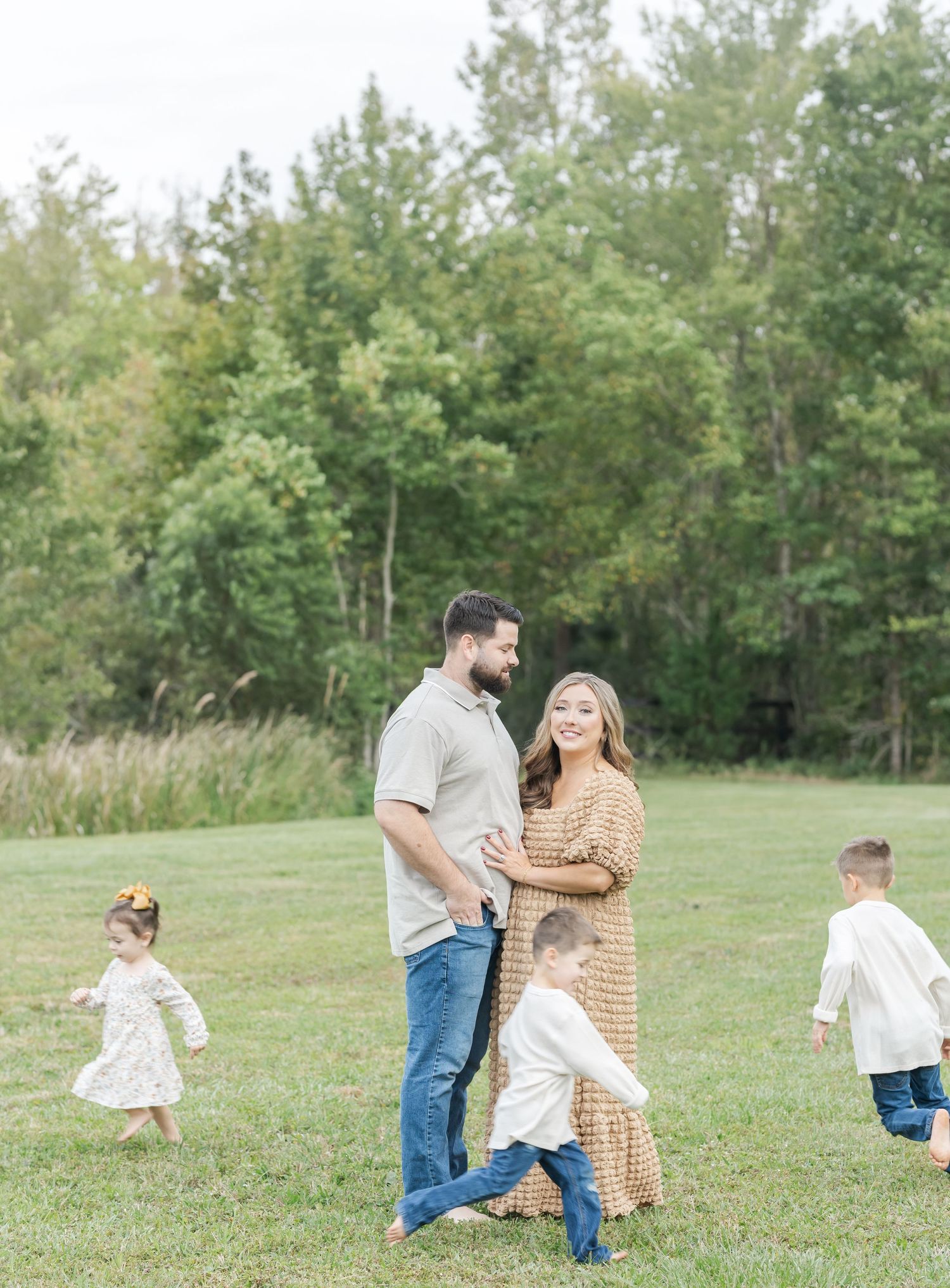 Family plays and runs together in a grassy field surrounded by trees on a sunny day.