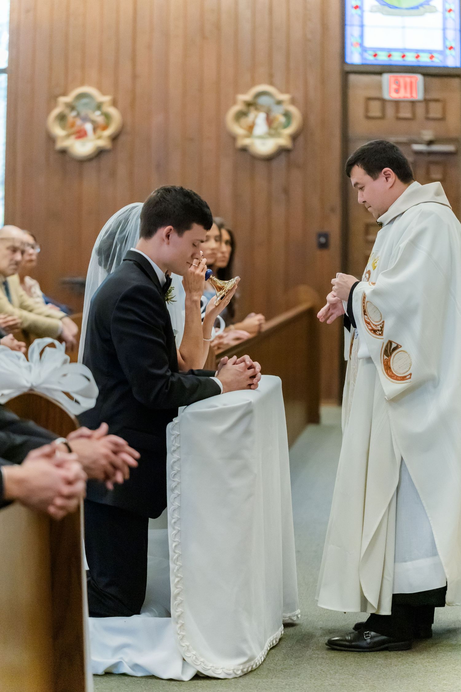 A wedding ceremony moment with a priest in white robes performing a blessing at the altar.