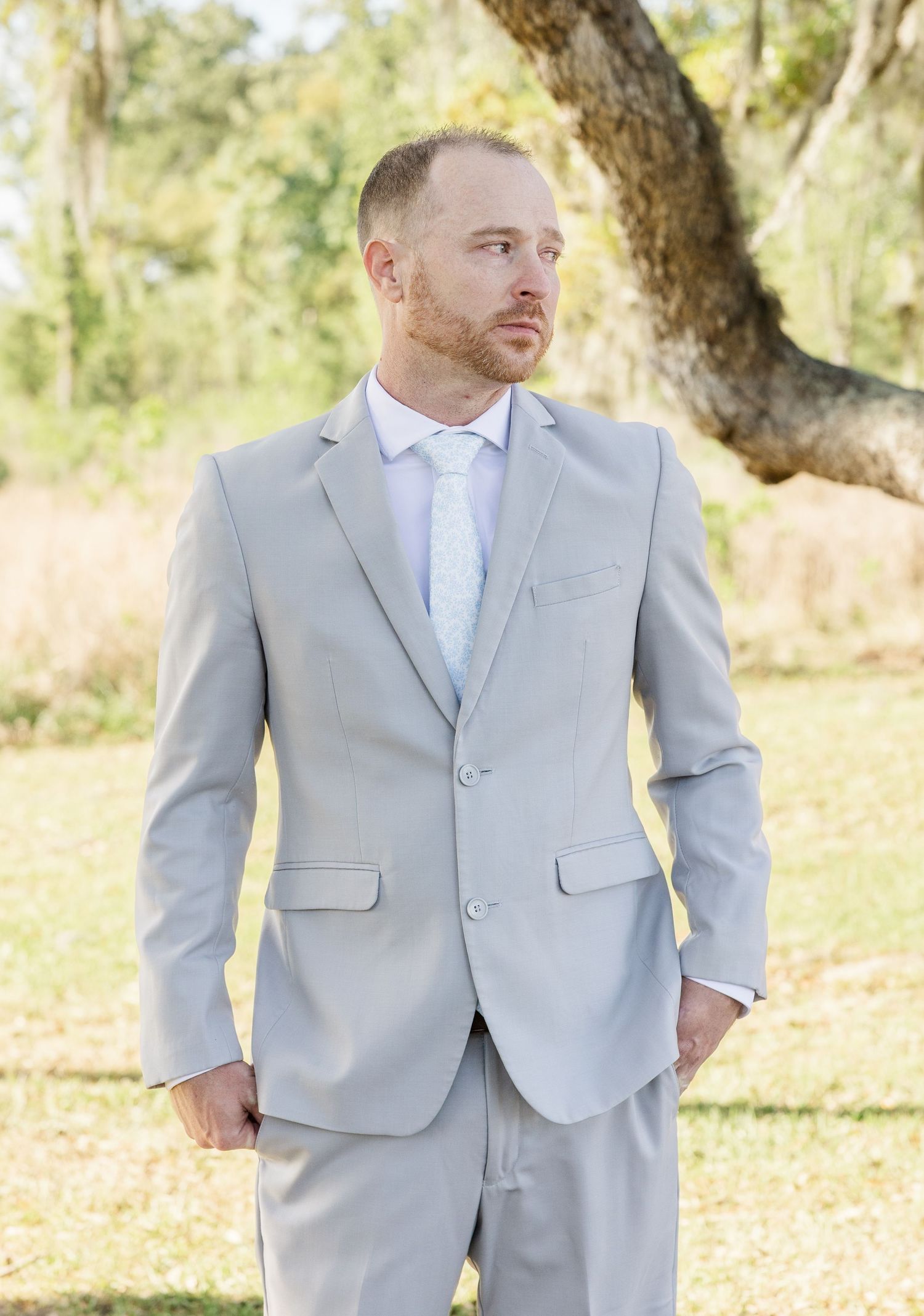 Groom in a light gray suit and tie standing outdoors near a tree on his wedding day.