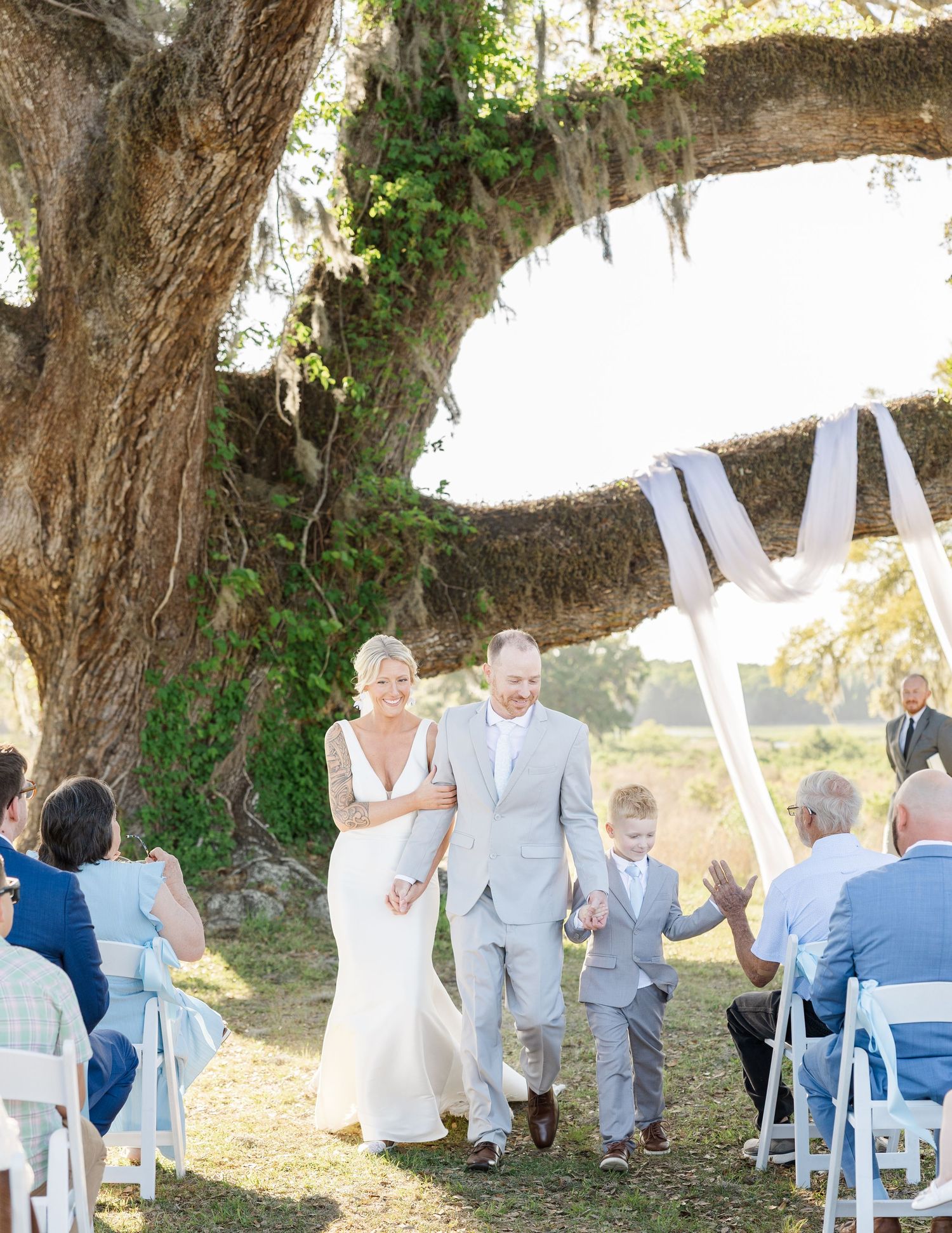 Bride and groom walk down outdoor aisle under a large oak tree draped with Spanish moss at a sunny wedding ceremony.